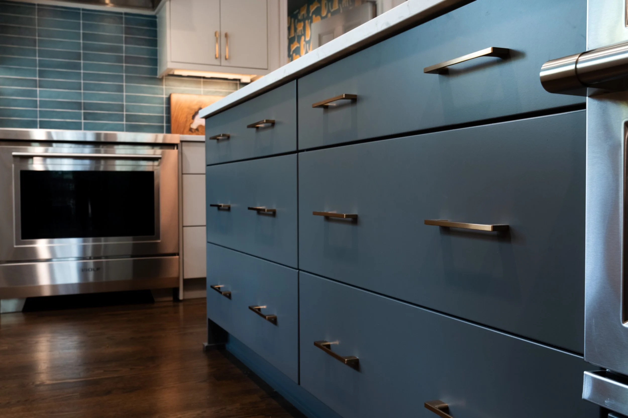 A modern kitchen with dark hardwood floors, blue cabinets with gold handles, a white marble countertop, teal subway tile backsplash, a stainless steel oven, and white upper cabinets. There is a colorful abstract painting on the wall behind a wooden cutting board.