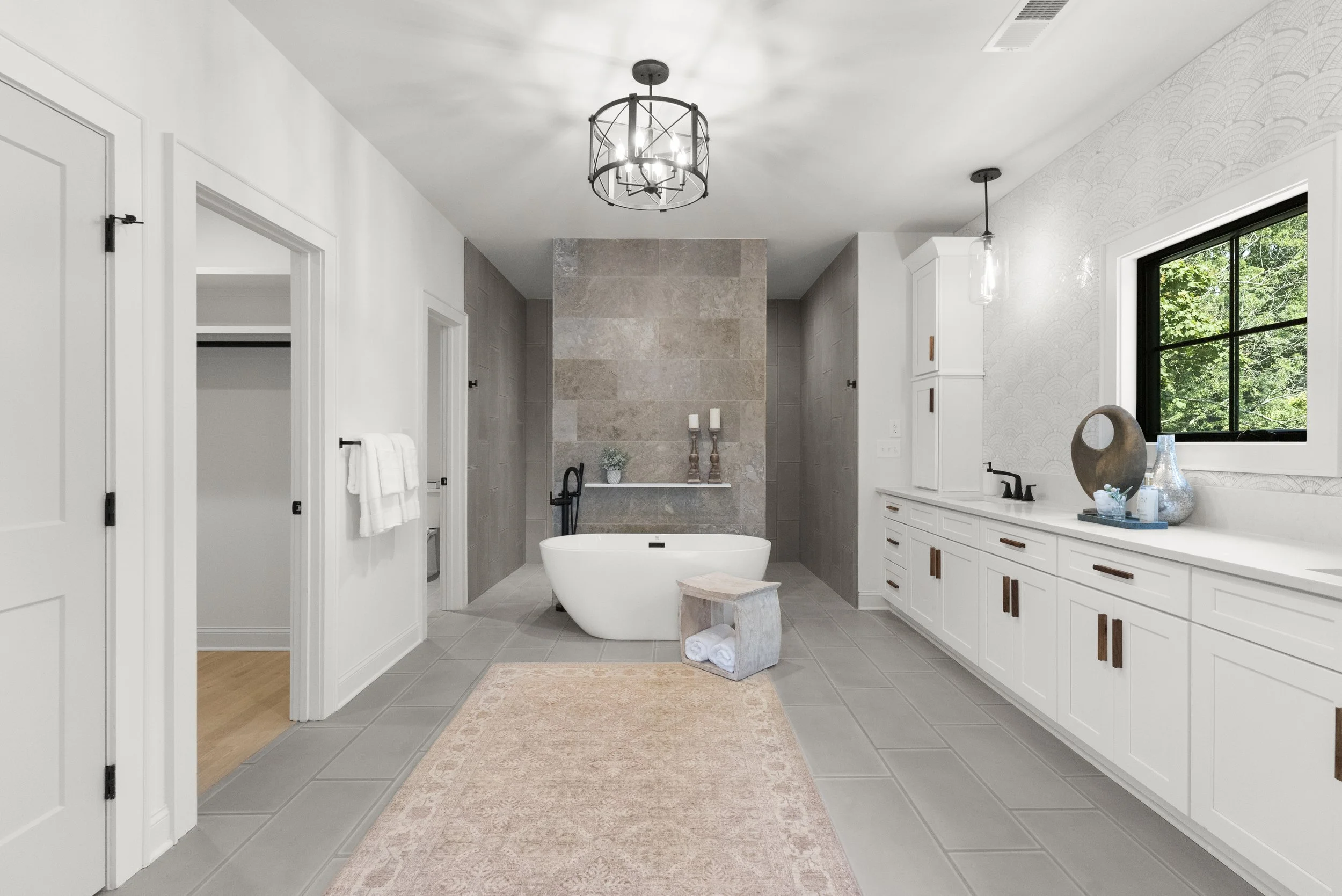Modern bathroom with a freestanding bathtub, white vanity cabinets, a wall of tiles, a window showing trees outside, a rug, vases, and decorative candles.