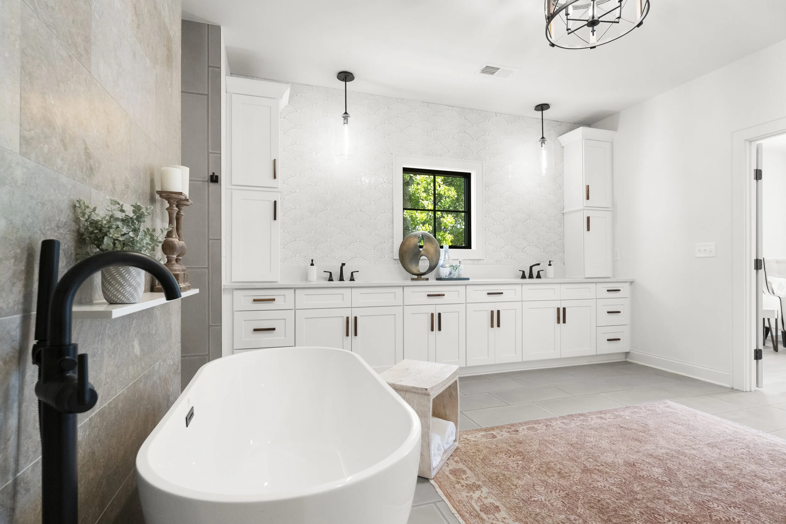 Modern bathroom with a freestanding bathtub, white cabinetry, a window with green trees outside, and pendant lighting.