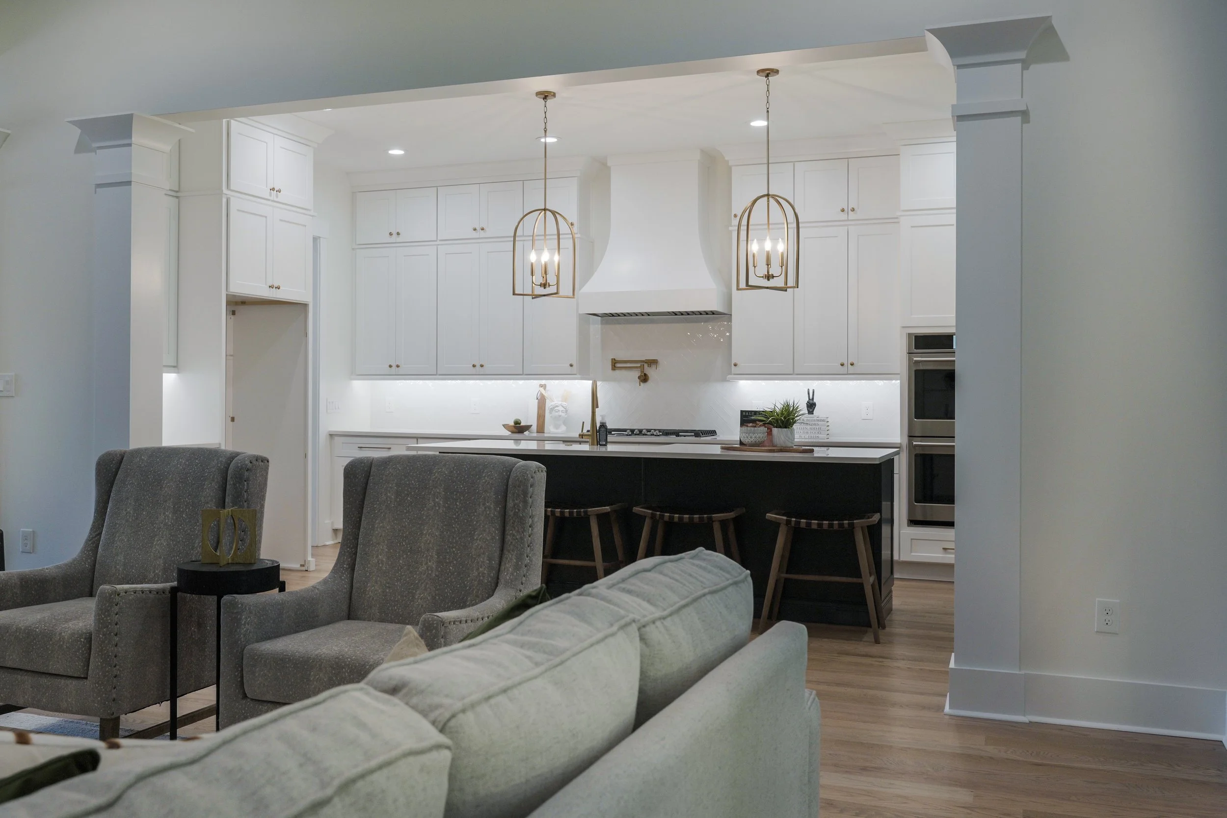 Modern open-concept kitchen featuring white cabinetry, pendant lighting, and a kitchen island with bar stools, adjacent to a living room with gray upholstered chairs.