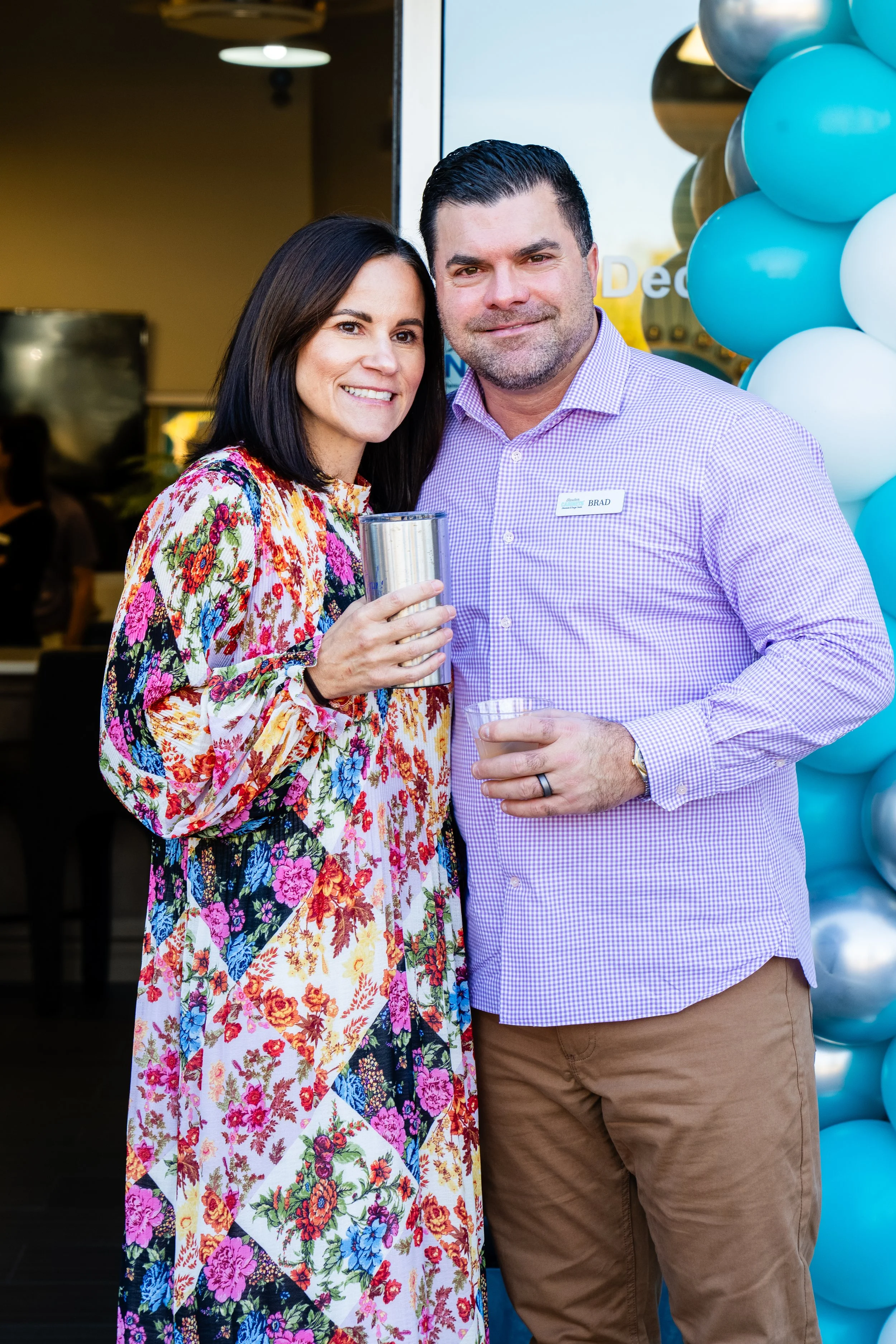 A woman with dark hair wearing a colorful floral dress, smiling and holding a drink, standing next to a man with dark hair and a beard, wearing a purple checkered shirt and brown pants, in front of blue and white balloons.
