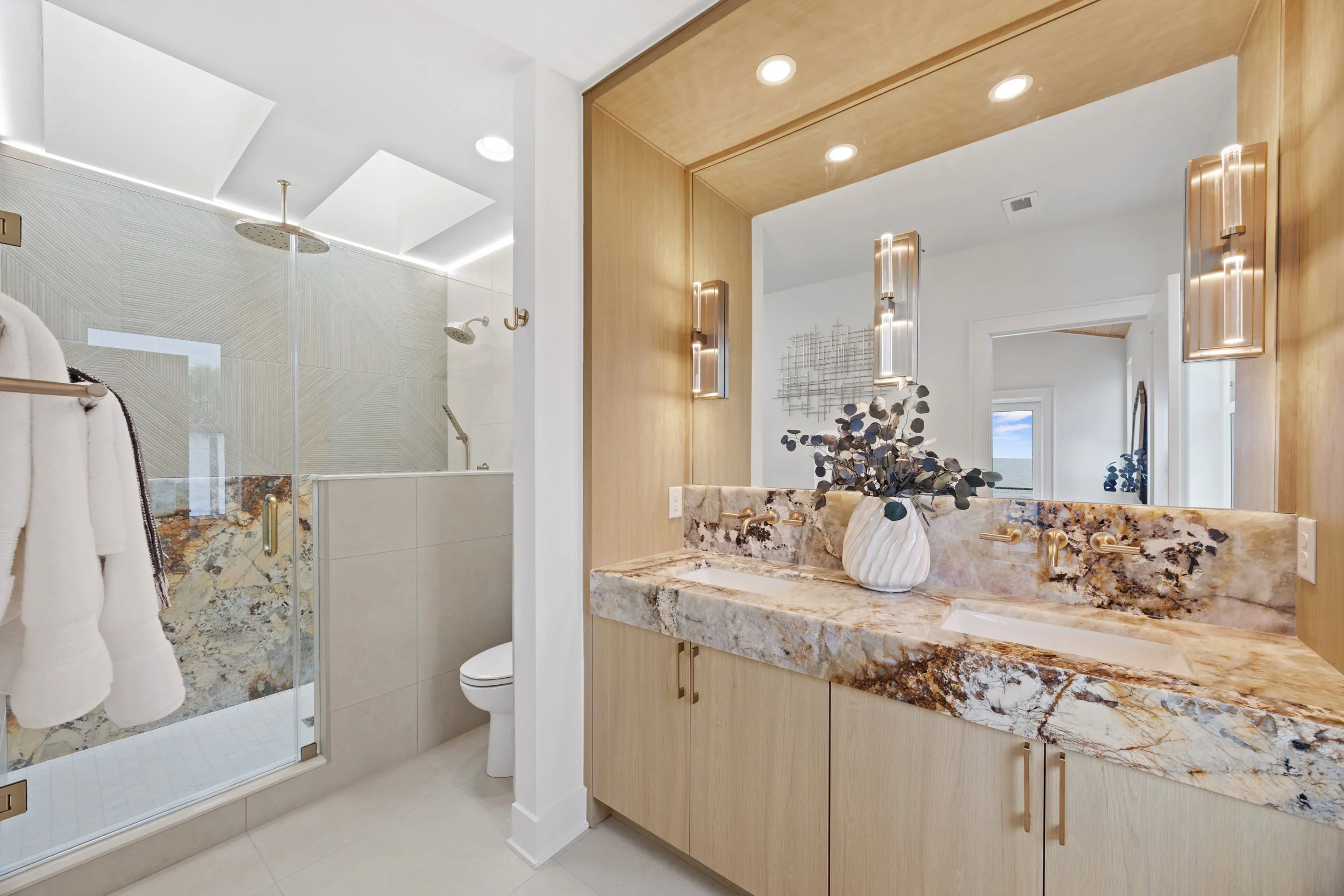 Modern bathroom with a glass-enclosed shower, a wooden vanity with a marble countertop, a large mirror, wall-mounted lights, and decorative elements including a vase with eucalyptus branches.