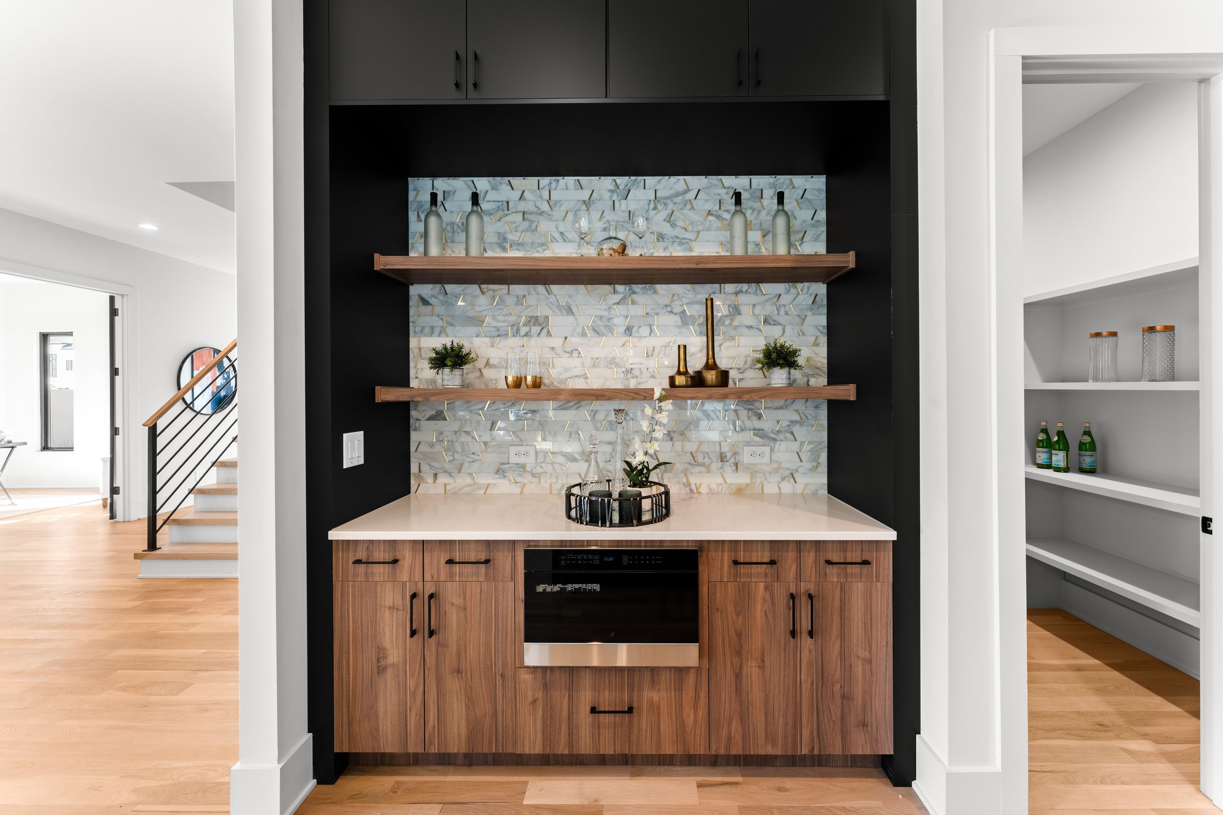 Kitchen area with a built-in wood cabinet, black oven, and black shelves decorated with bottles, jewelry, and greenery on a marble backsplash.
