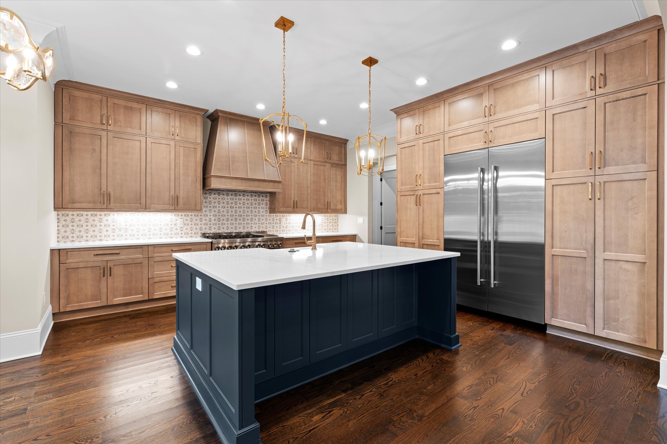 Modern kitchen with wooden cabinets, a central island painted navy blue, white countertops, a stainless steel refrigerator, and a patterned backsplash, illuminated by ceiling lights and pendant lights.