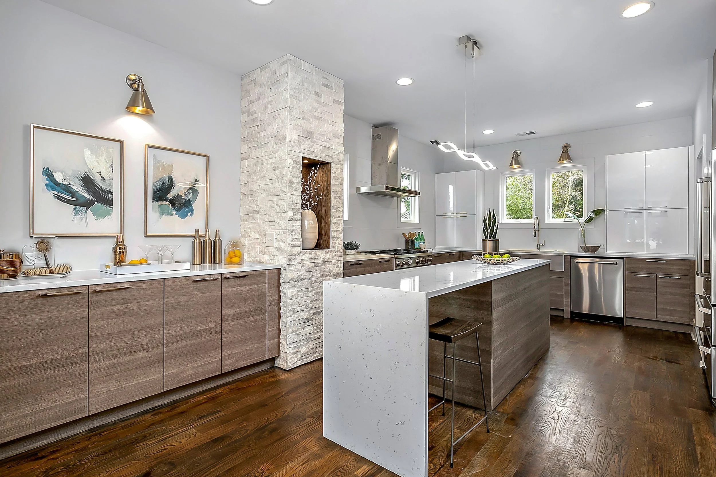 Modern kitchen with white walls, wooden cabinets, a large white island, and stainless steel appliances. Artworks and decorative bottles are on the counter, with plants near the windows and pendant lights hanging from the ceiling.