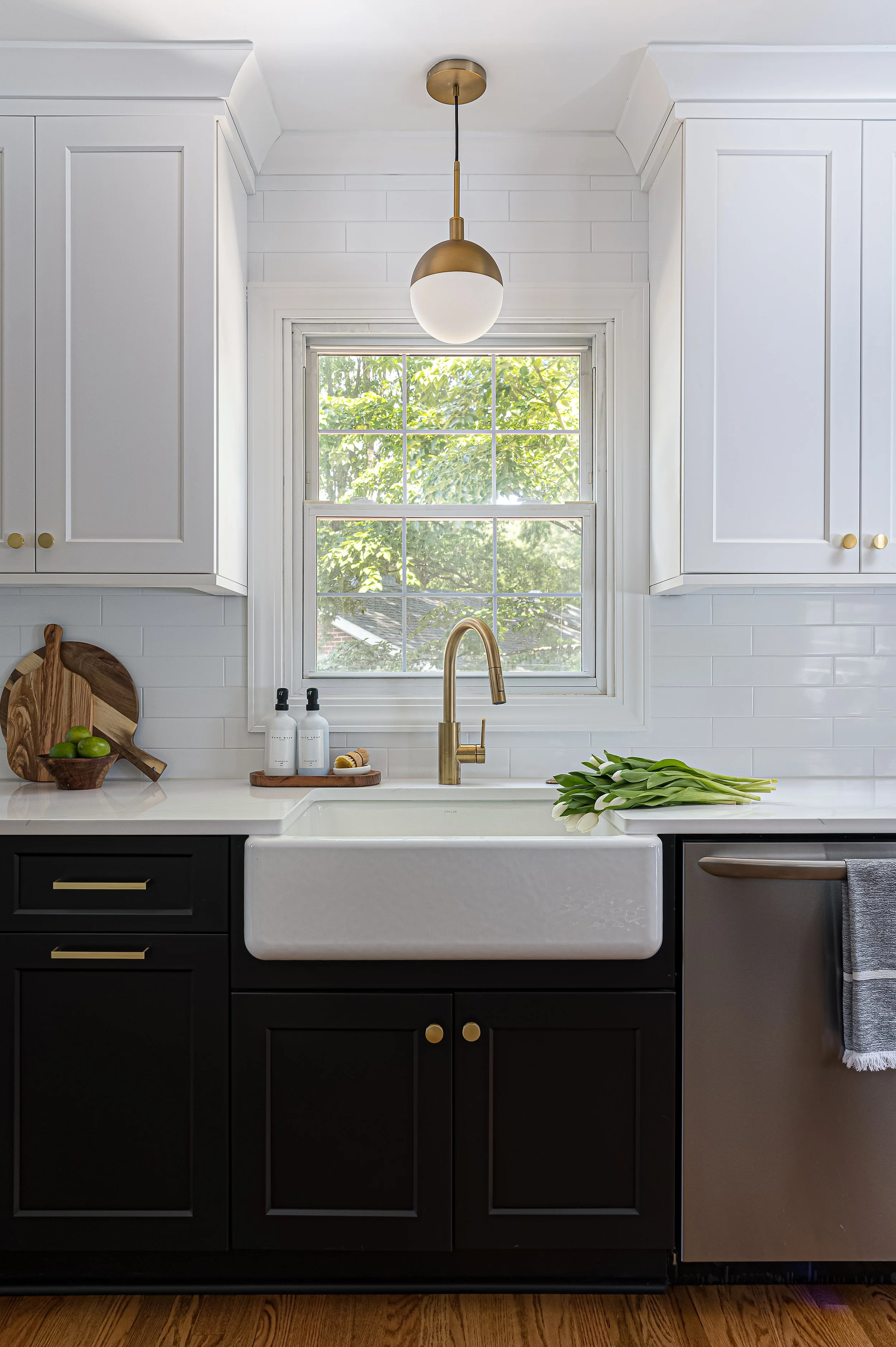Kitchen with white upper cabinets, black lower cabinets, a farmhouse sink, a window with greenery outside, a wooden chopping board, soap bottles, a bundle of tulips on the countertop, a drawer towel, and a gold pendant light hanging above the sink.