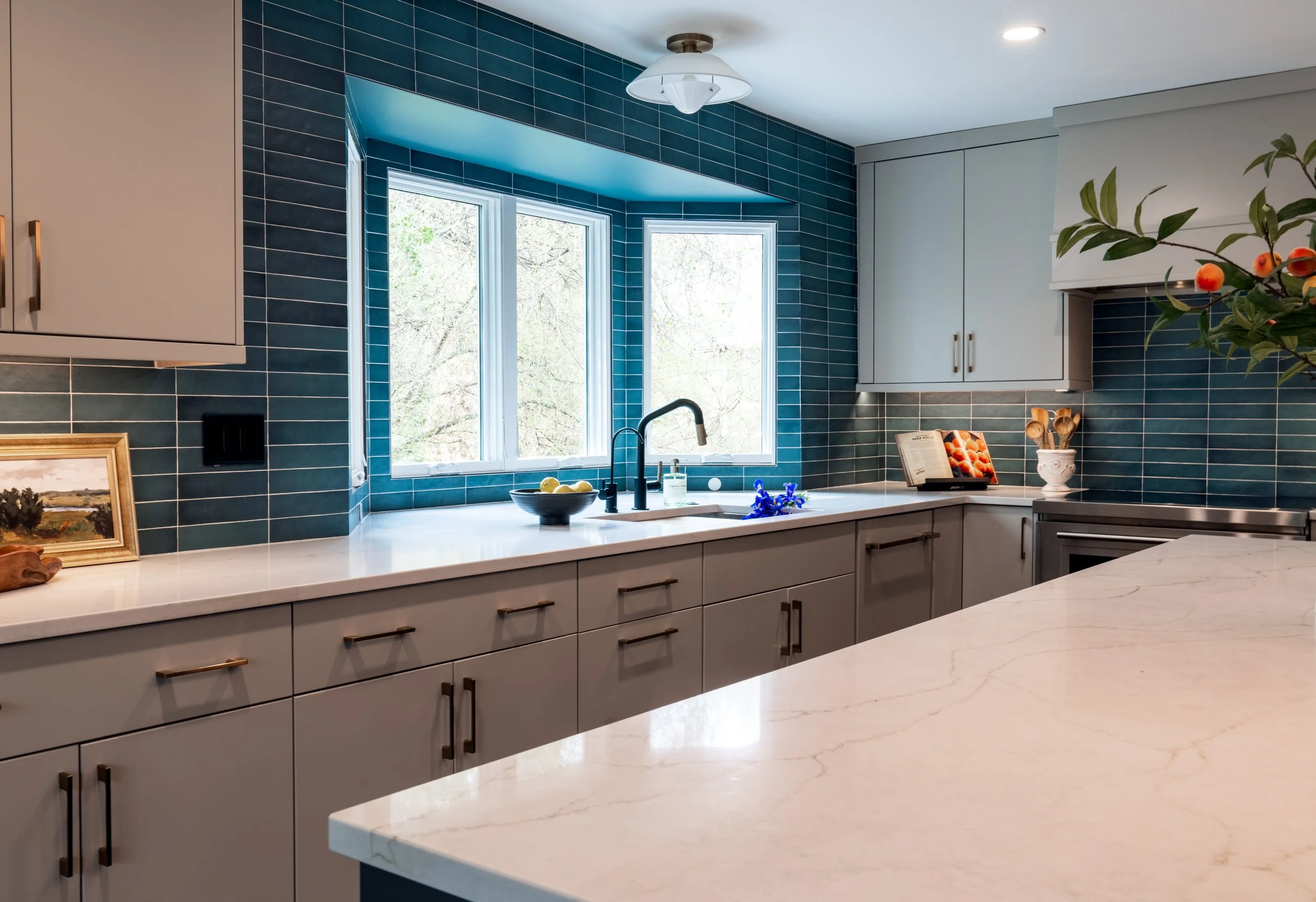 Modern kitchen with white cabinets, dark blue tiled walls, and a large window above the sink overlooking trees. There are decorative items on the countertop, including a bowl of lemons, a book, and a potted plant with red berries.