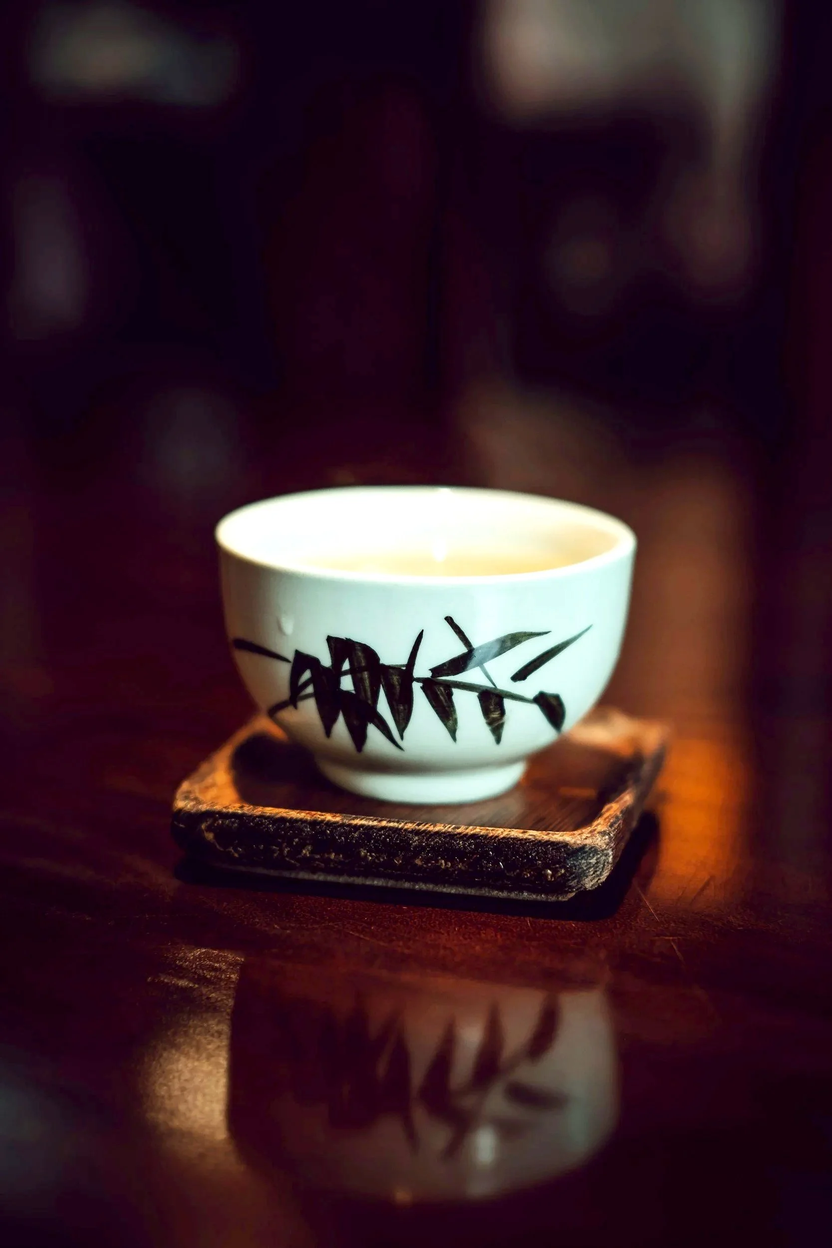 A white ceramic teacup with black bamboo leaf design, placed on a small wooden tray on a dark polished wooden surface.