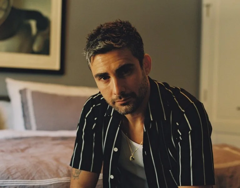 A man with short dark hair and a beard is sitting on a bed, looking at the camera, wearing a black shirt with white stripes over a white T-shirt, in a dimly lit room.