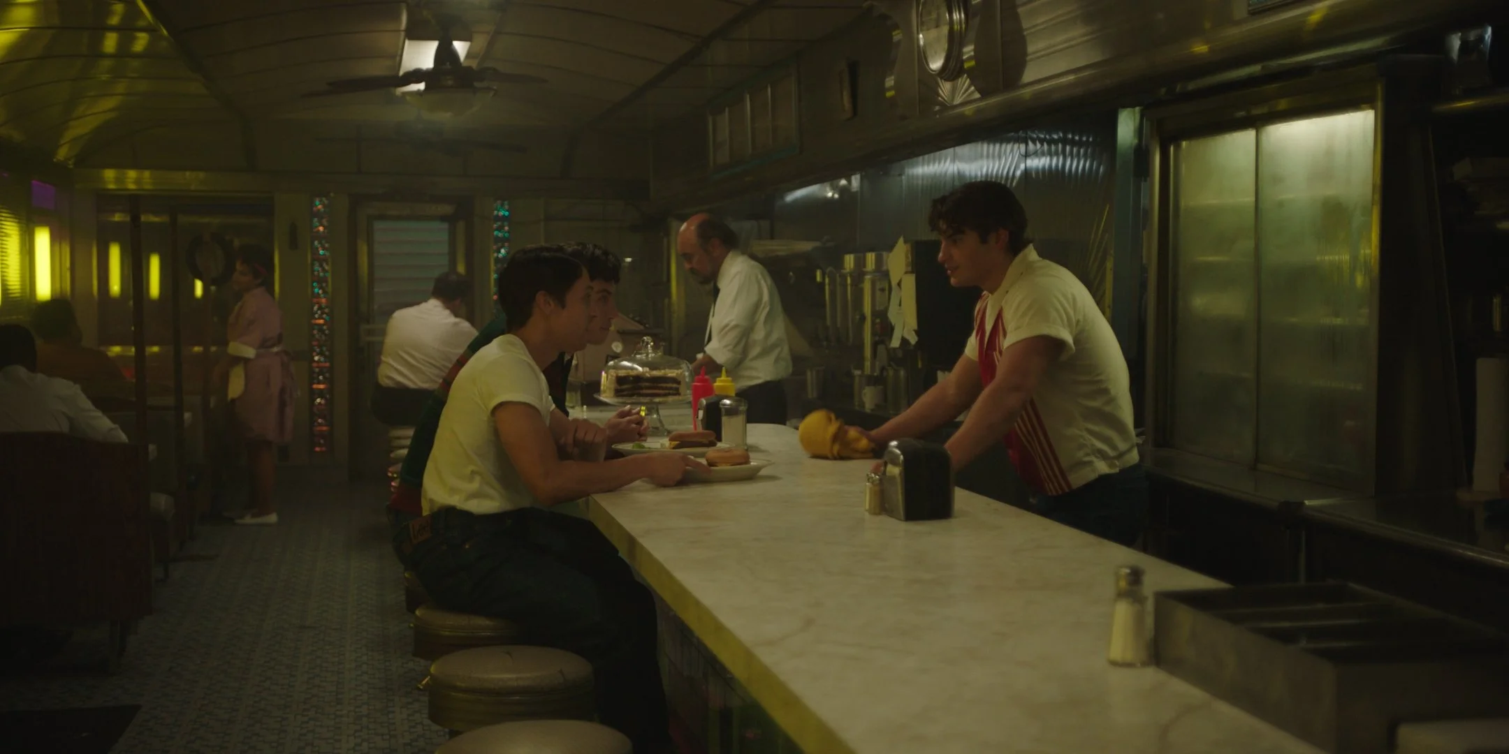 People sitting at a marble counter in a diner, with a chef preparing food behind the counter. The diner has a vintage feel with stained glass windows and a ceiling fan.