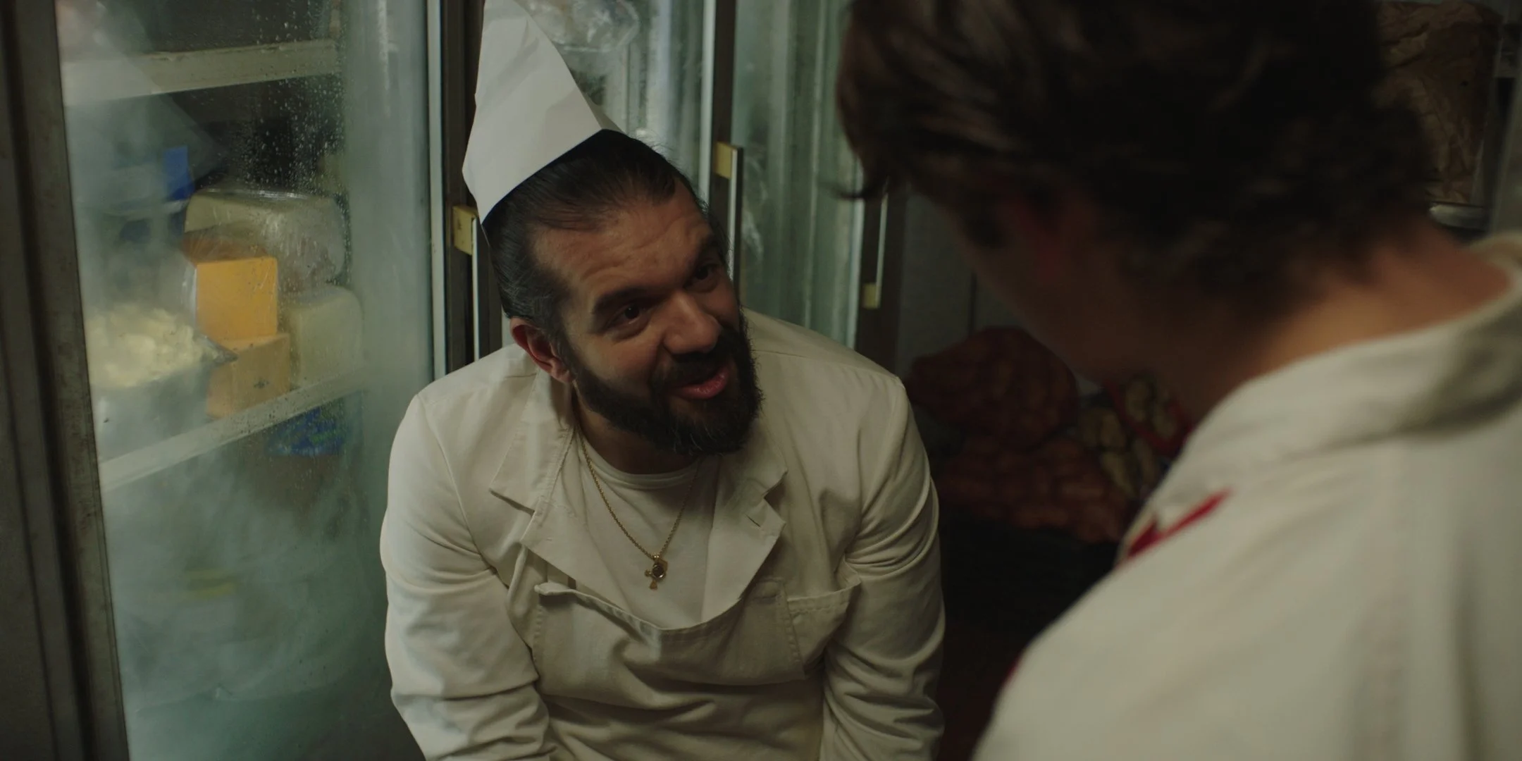 Two men, one dressed as a chef with a white coat or apron, a white hat, and a gold necklace, having a heated conversation in a kitchen. The other man is partially visible from behind, wearing a white shirt with red markings. The scene appears intense
