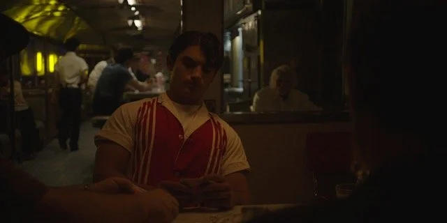 A young person sitting at a table in a dimly lit restaurant, playing a card game, with other patrons and staff visible in the background.