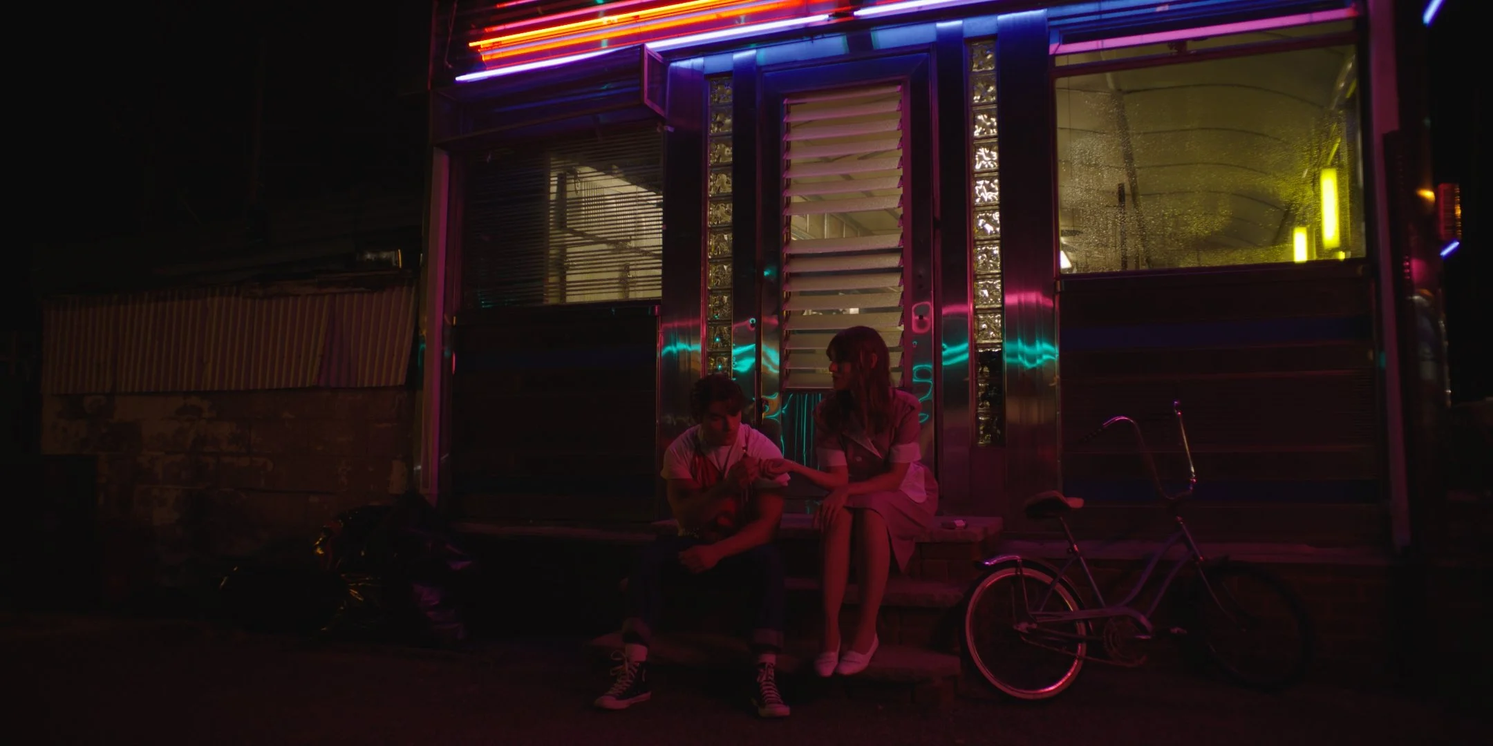 A person with short hair and a person with long hair sitting on the steps outside a store with neon lights at night, with a bicycle parked nearby.