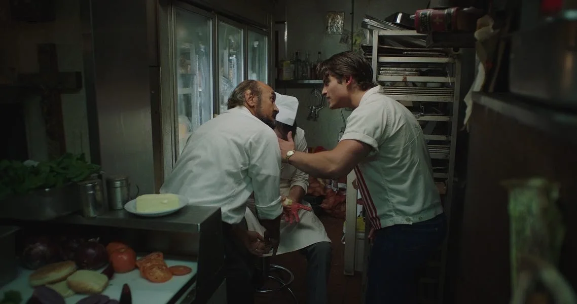 A tense confrontation in a small kitchen, with two men, one older with a beard and the other younger, facing each other closely. The older man is seated and the younger is leaning forward with a hand on the older man's shoulder. There is a third pers