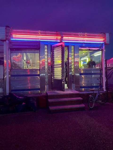 A small illuminated food or vendor stand with neon lights in purple, red, and yellow, featuring open doors revealing the interior with people inside, set outdoors at night, with a bicycle parked outside.