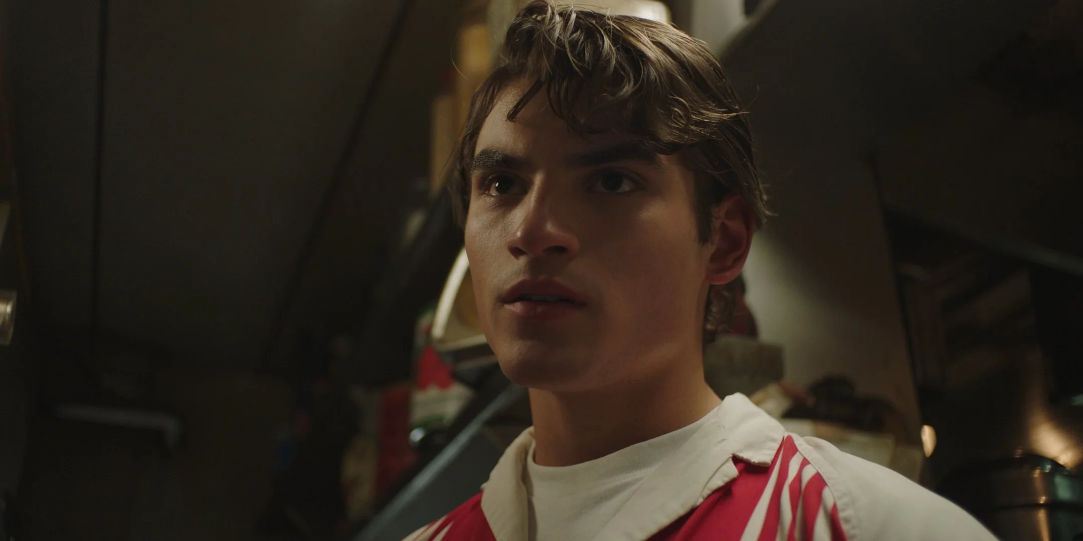A young man with curly brown hair and wearing a white shirt with a red jacket, standing in a dimly lit kitchen.