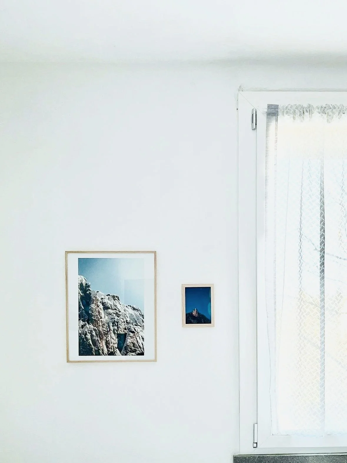 Due stampe fotografie di paesaggi montani realizzate da Martina Chiarato - thatsmarch appese a una parete bianca accanto a una finestra con tende leggere.