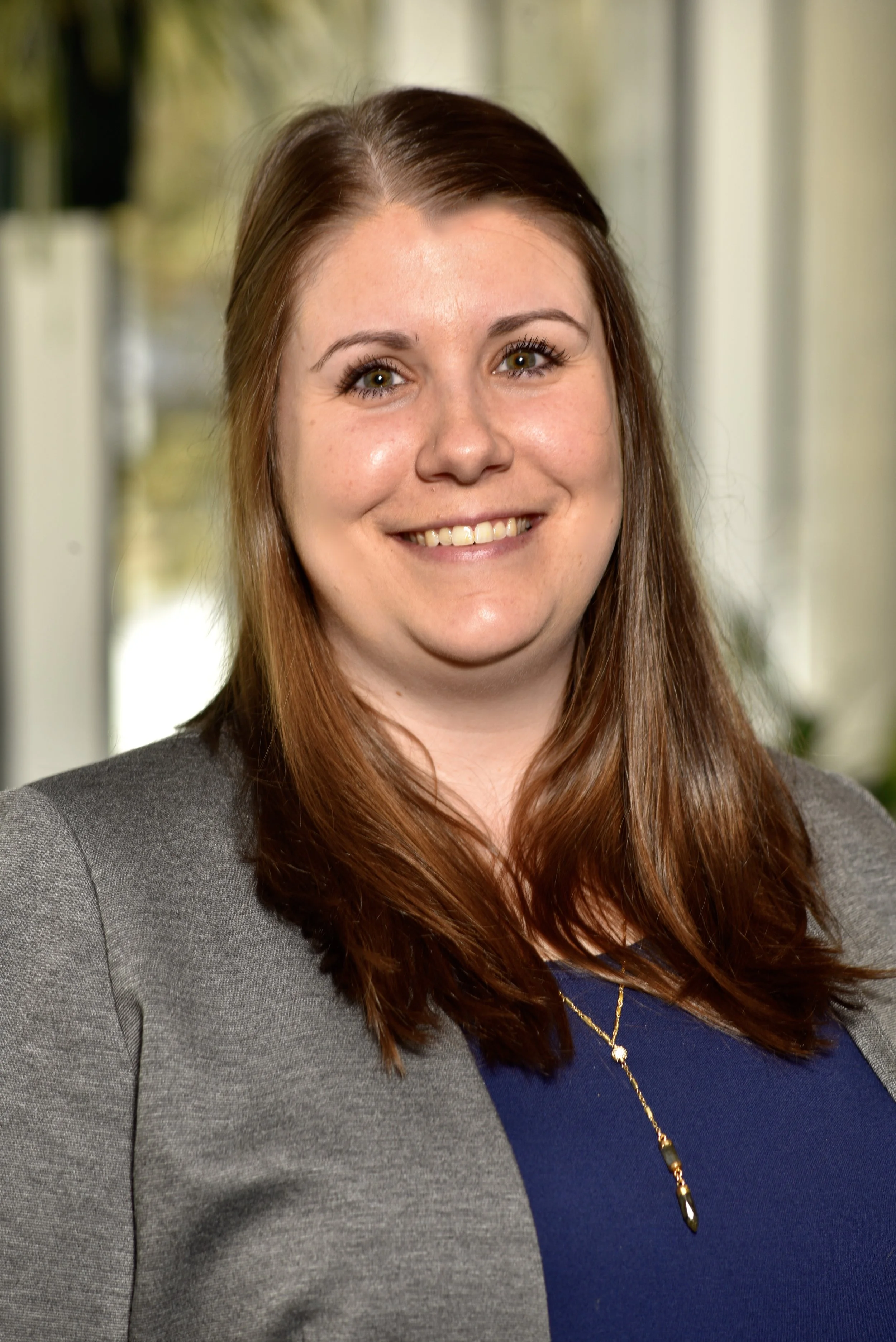 A picture of Dr. Kirsten Gullickson, a smiling woman with long brown hair. She is wearing a gray blazer over a dark blue top with a gold necklace. She is standing indoors with a blurred background of windows and greenery.
