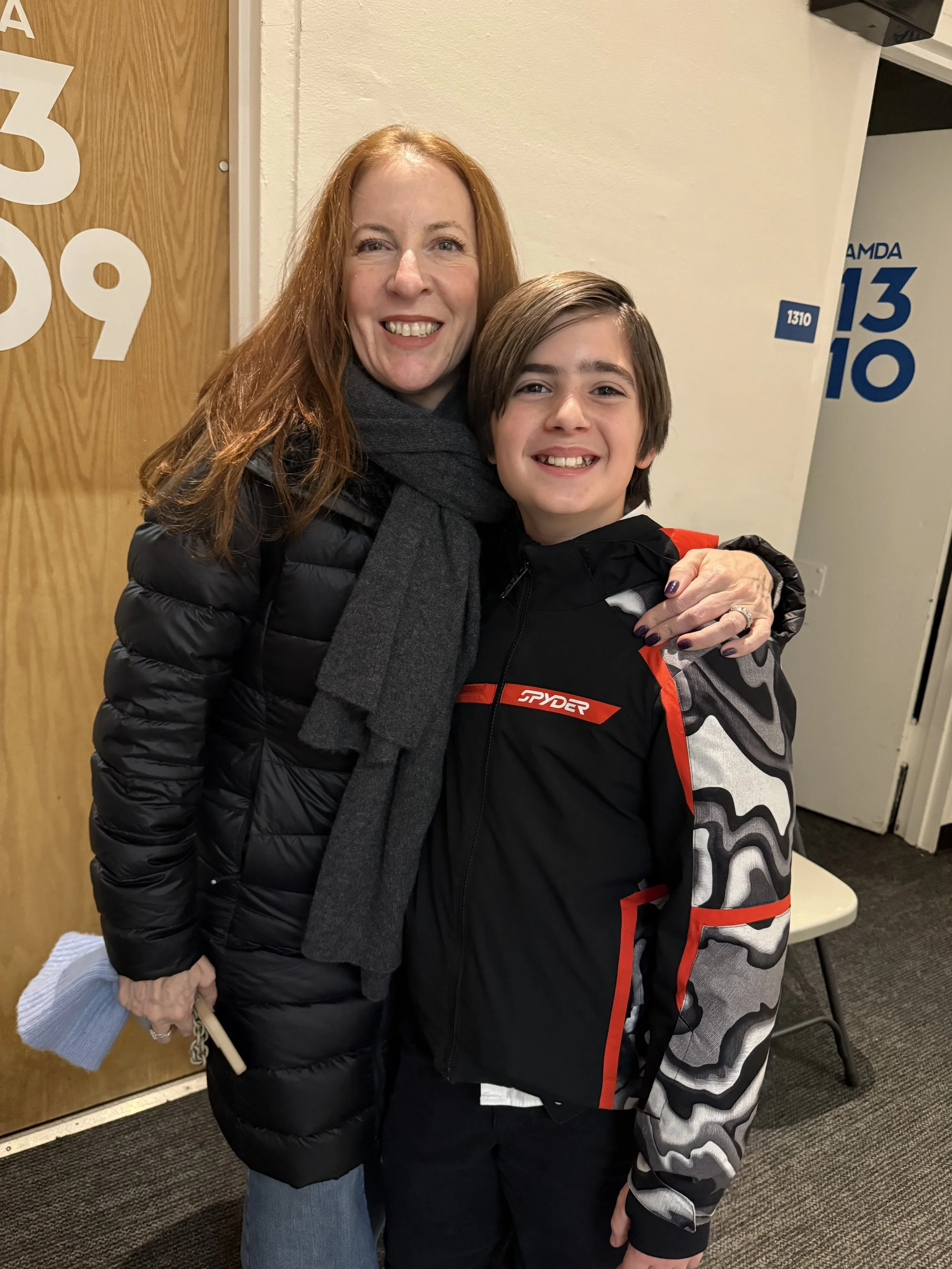A woman with long red hair wearing a black puffer jacket and gray scarf hugging a young boy with medium length brown hair wearing a black and gray camouflage jacket with red accents, smiling at the camera indoors.