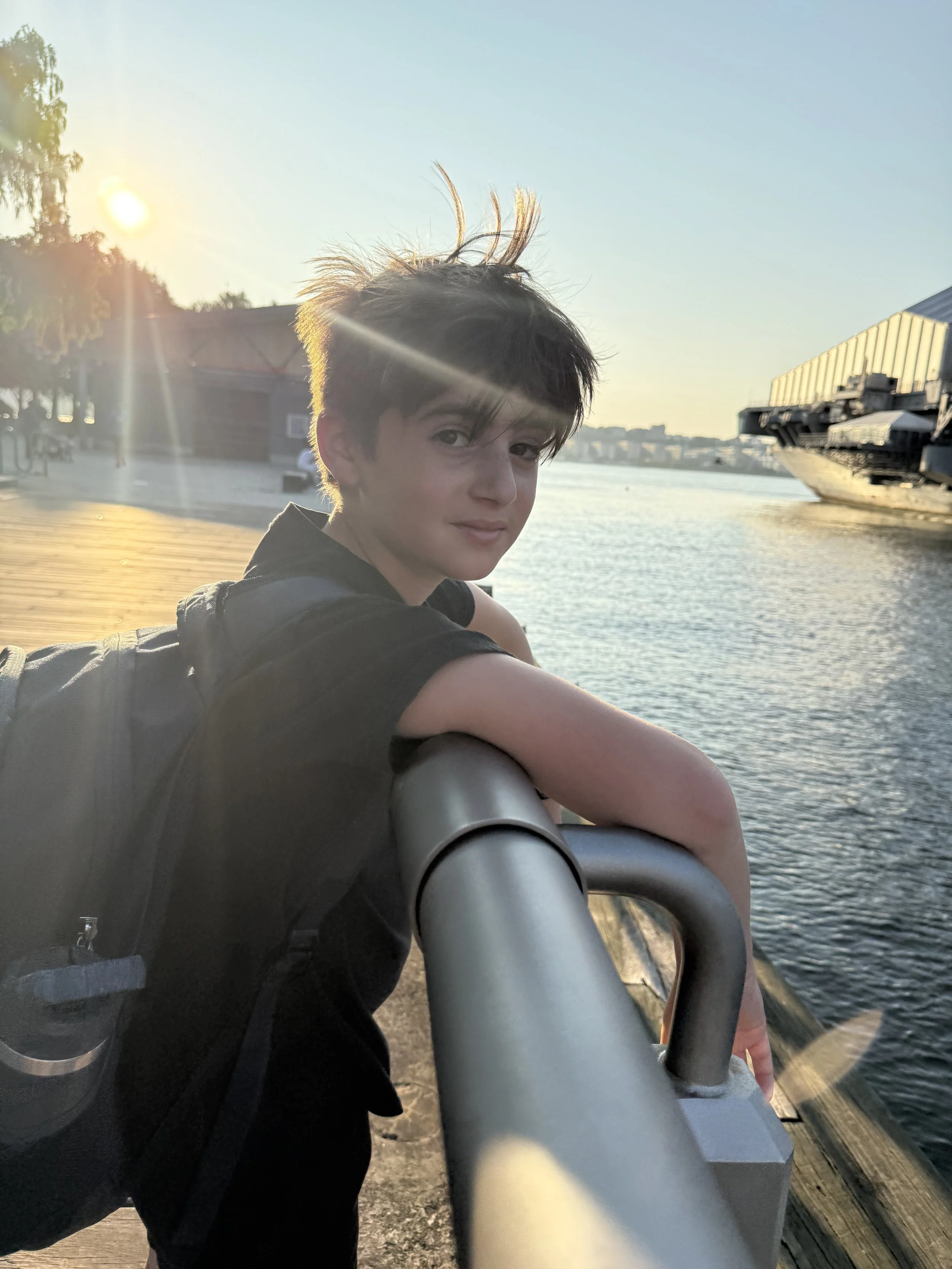 A young person with messy brown hair leans on a metal railing by the water during sunset, with industrial buildings and a cargo ship in the background.