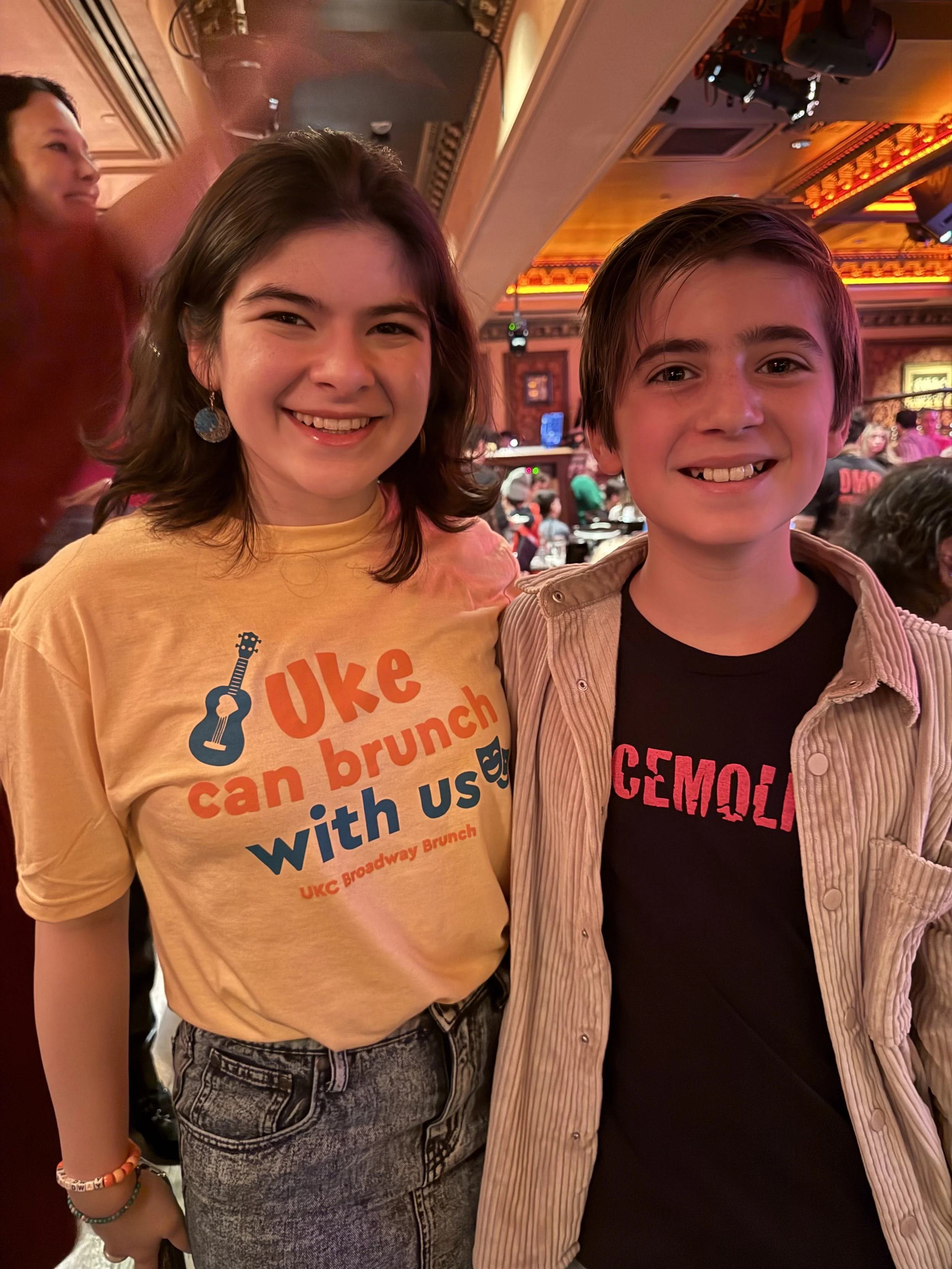 Two smiling children posing together at an event or celebration in a decorated venue with warm lighting and a crowd in the background.