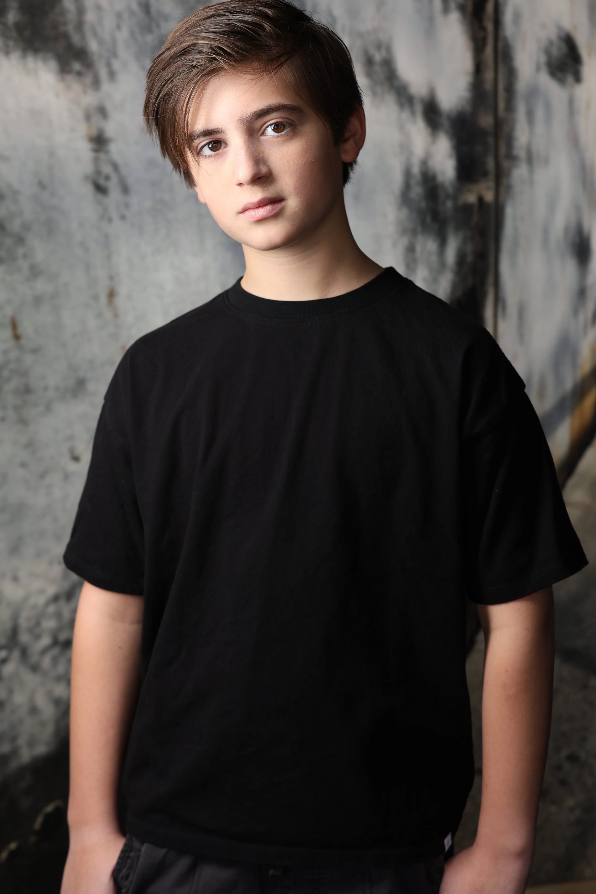 A young boy with brown hair and fair skin wearing a black t-shirt, standing against a textured, weathered wall background.