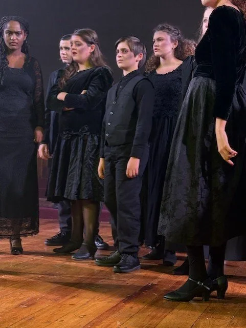 Group of children standing on a stage, dressed in black, possibly performing or rehearsing.