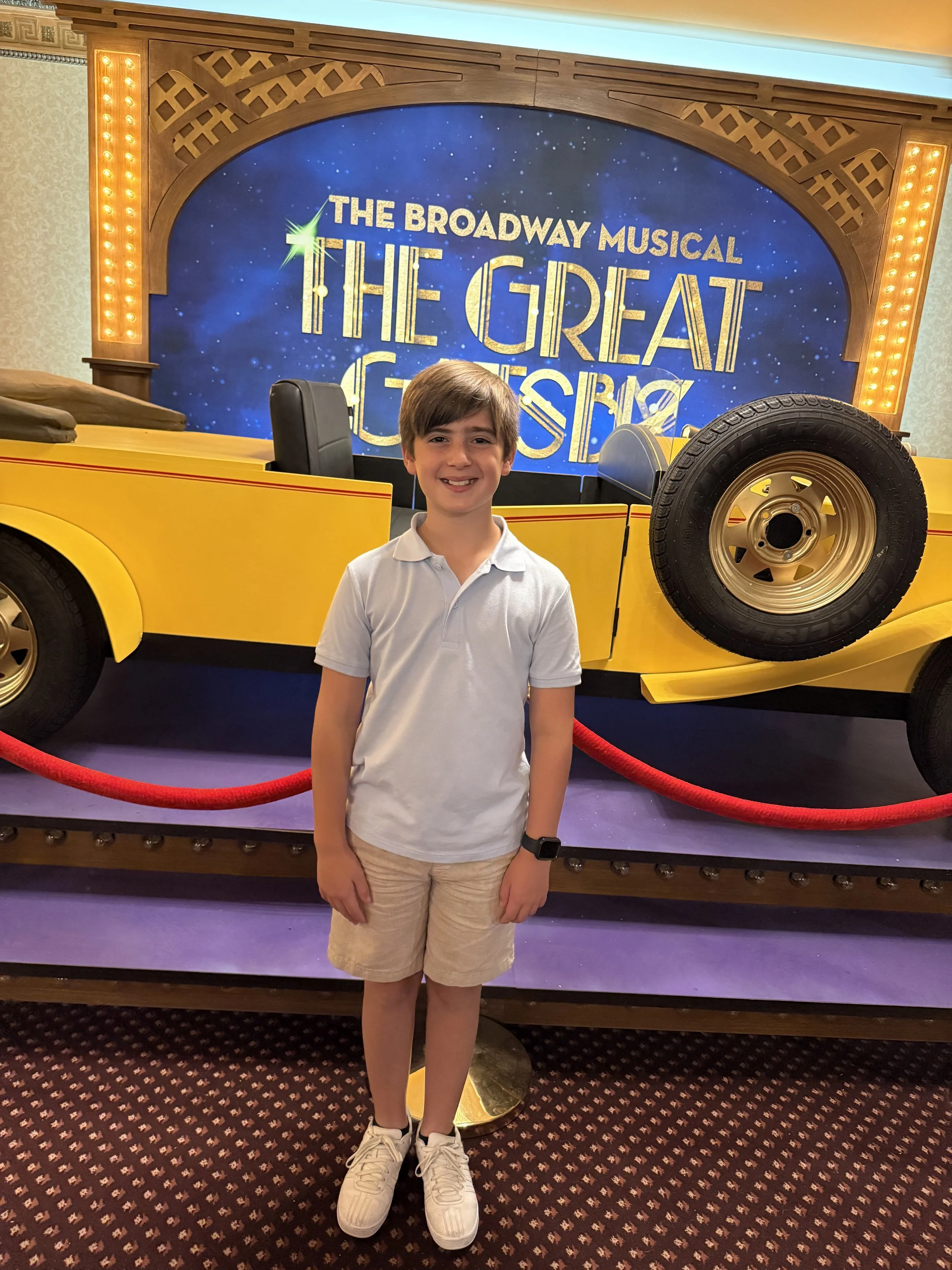 A young boy has brown hair and is smiling while standing in front of a yellow vintage car display at a Broadway musical event. The backdrop features a blue space-themed background with the text 'The Broadway Musical The Great Gats...'