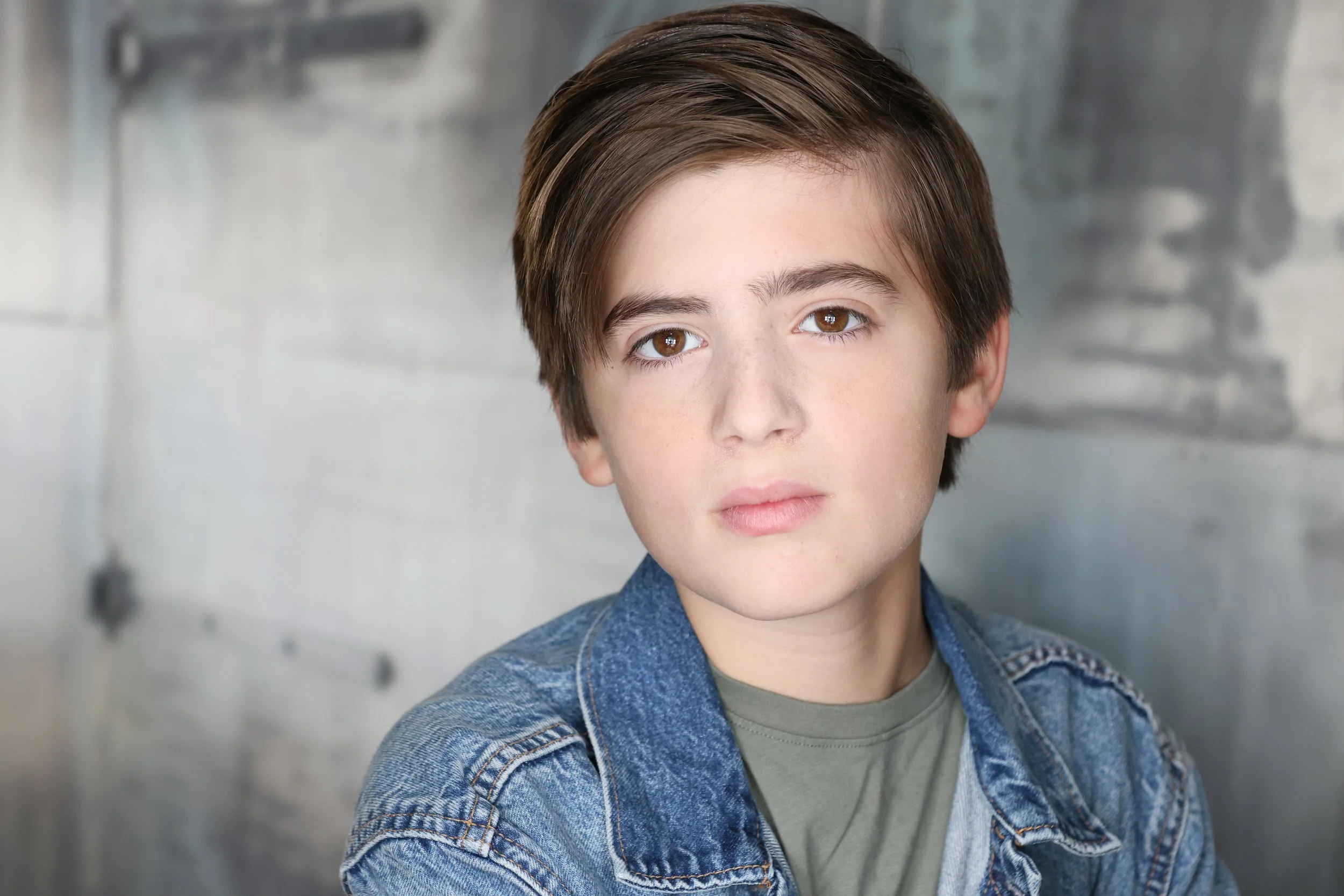 Close-up of a young boy with brown hair and brown eyes, wearing a denim jacket and gray shirt, against a blurred gray wall background.