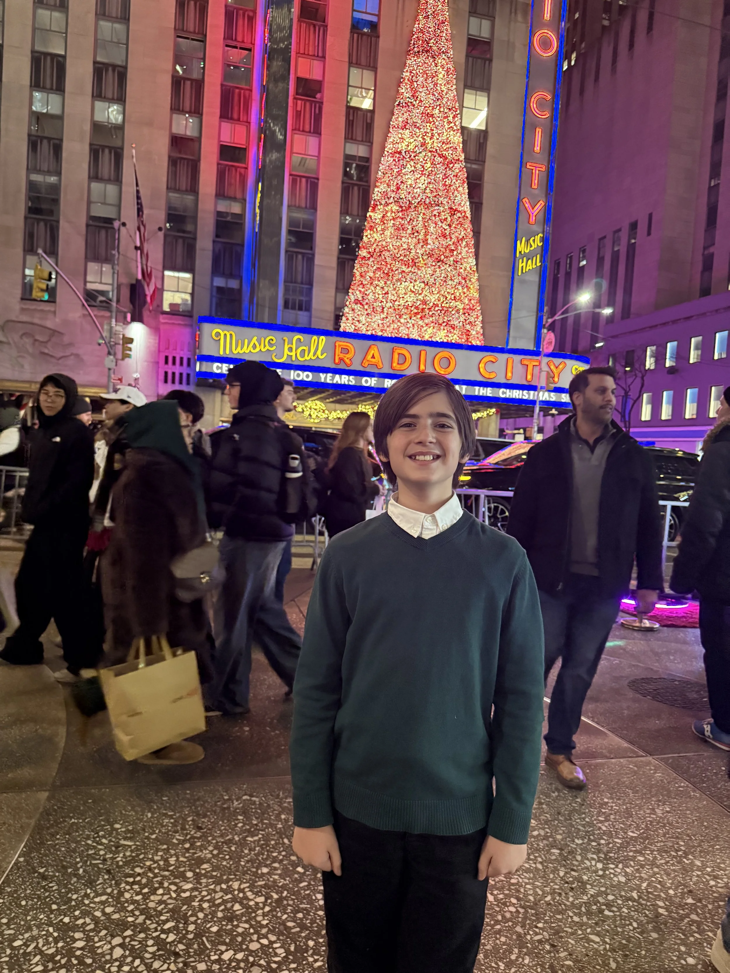 A boy smiling at night in Times Square, New York City, with a large illuminated Christmas tree and a Radio City Music Hall sign in the background.