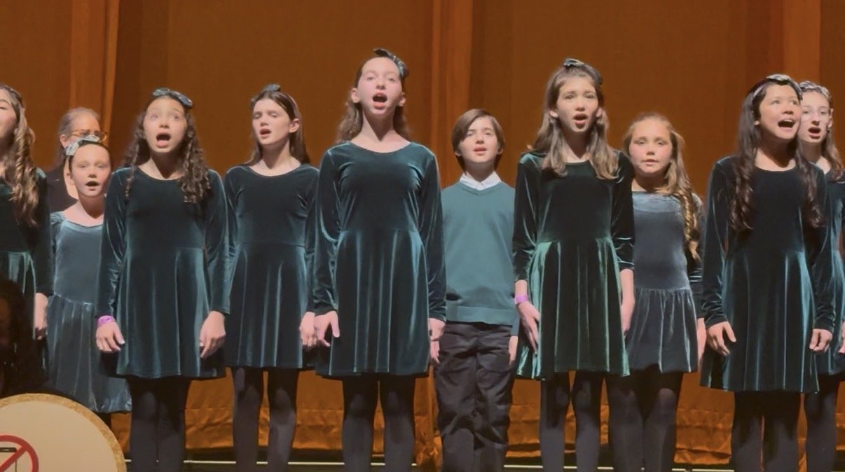A group of children singing on stage during a choir performance, dressed in matching dark green dresses and sweatshirts, with a wooden stage background.