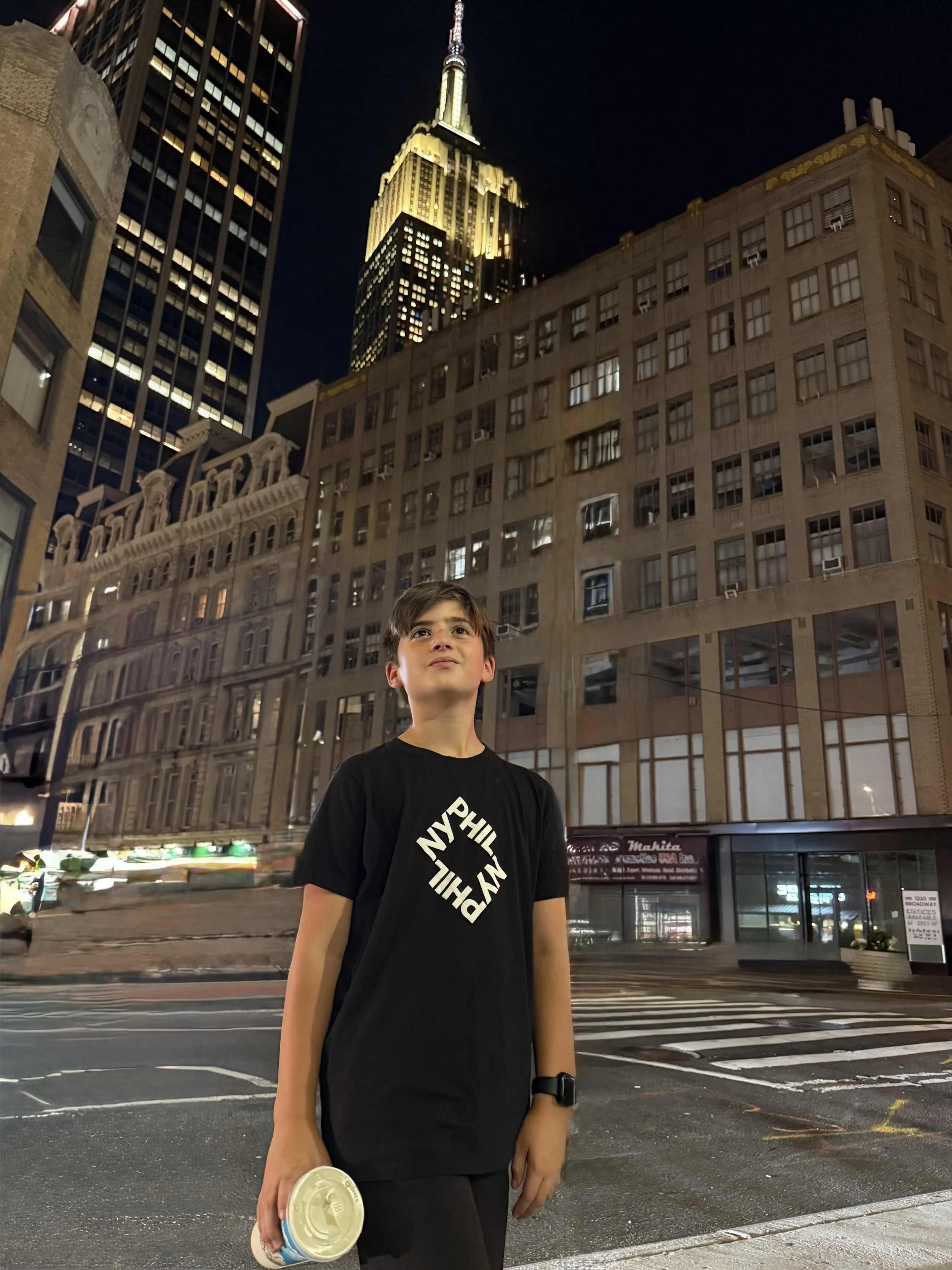 A young boy standing on a city street at night, holding a container, with tall illuminated buildings, including the Empire State Building, in the background.