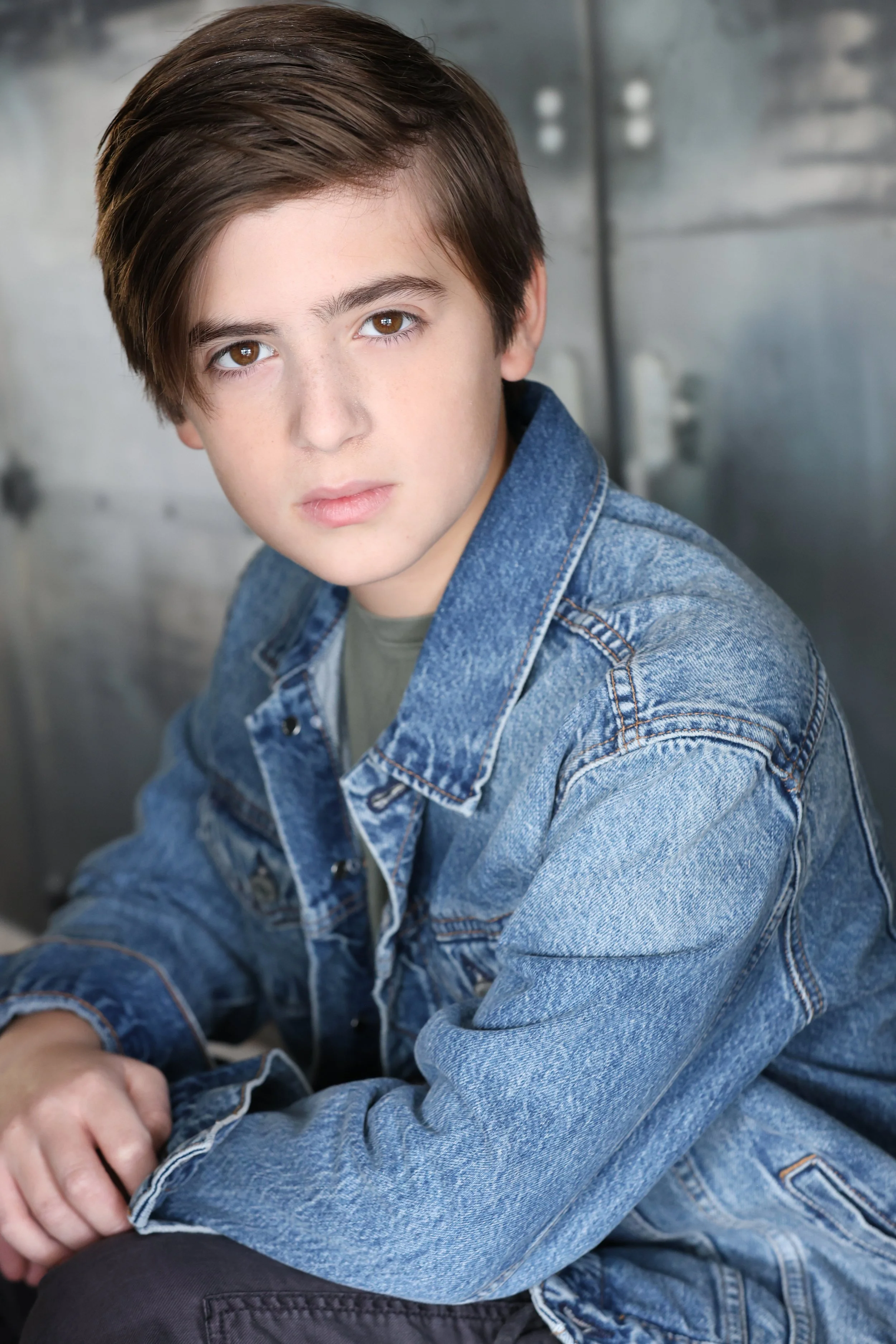 A young boy with brown hair and brown eyes wearing a denim jacket, sitting against a metallic background, looking at the camera with a serious expression.