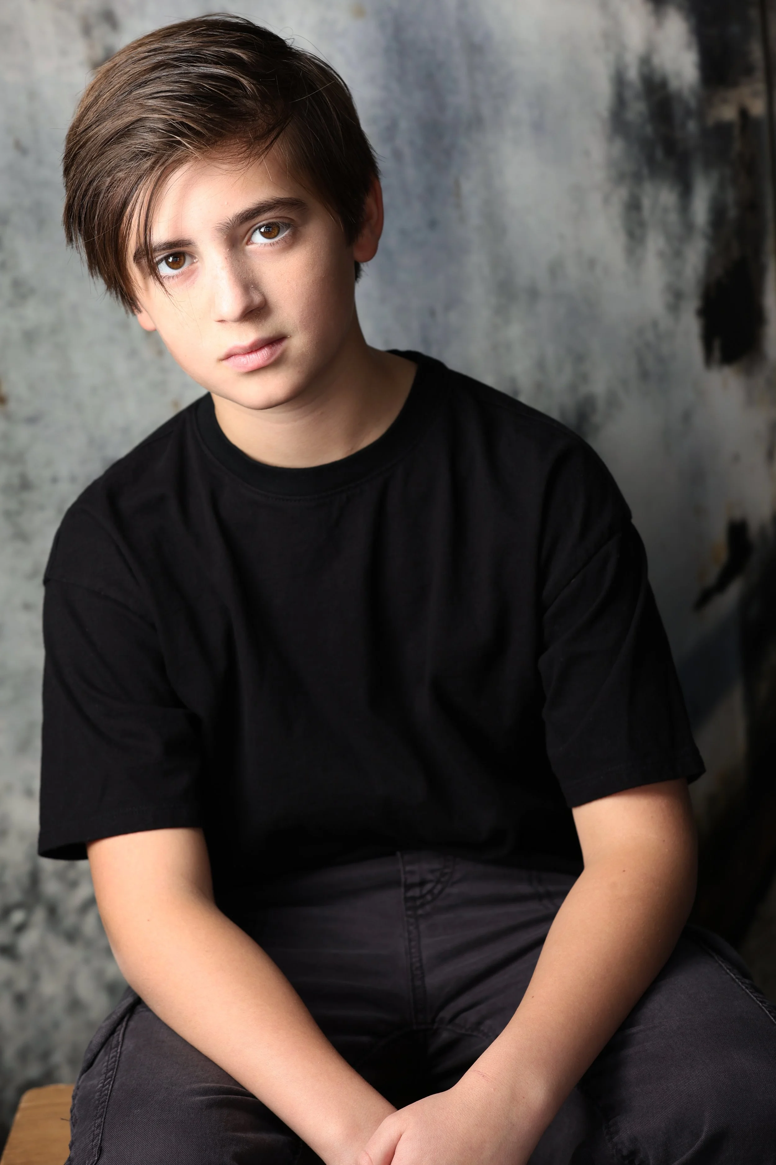 A young boy with brown hair and brown eyes, wearing a black t-shirt, sitting against a textured gray background.