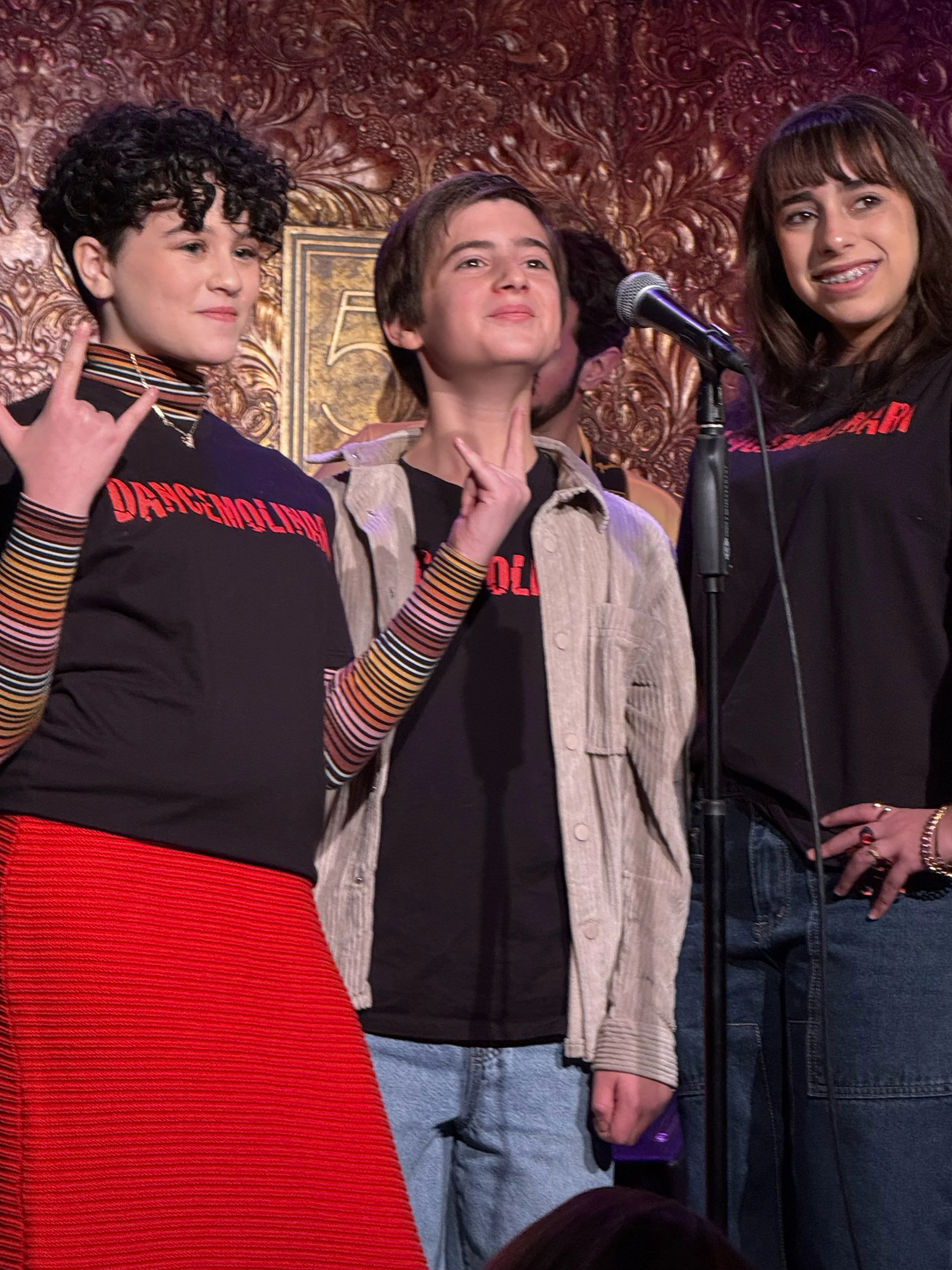 Three young women standing on stage in front of a textured, ornate golden wall, with microphone in front of them. They are making peace signs and have confident expressions.