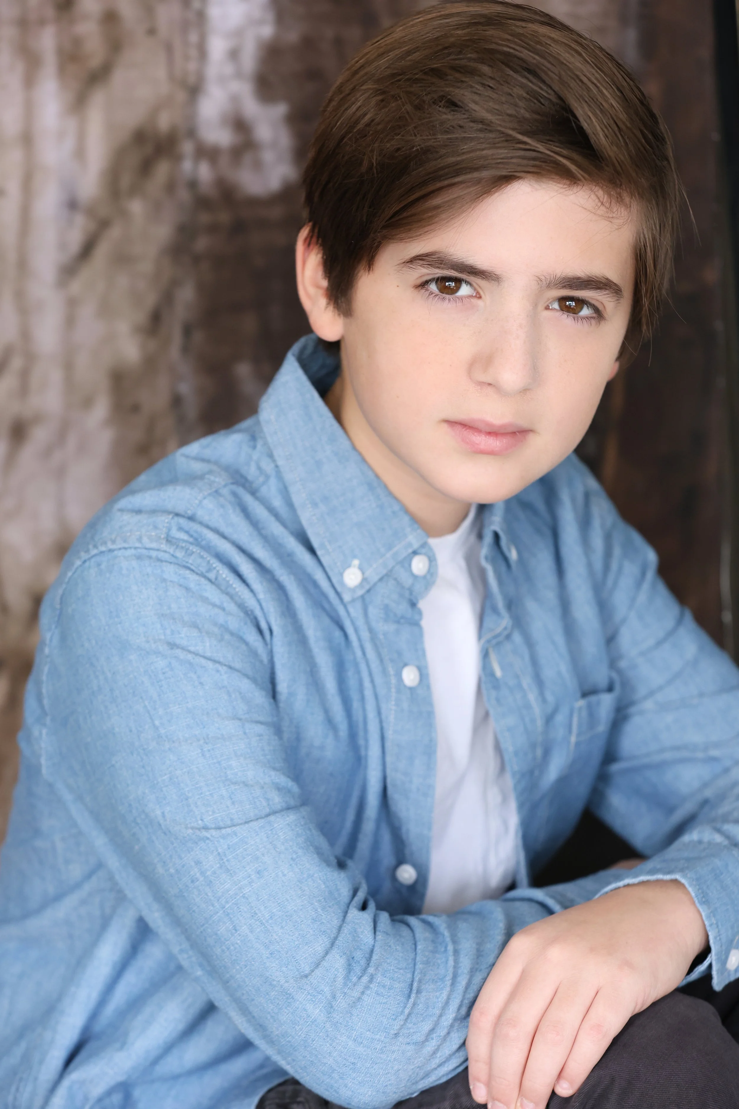 A young boy with brown hair and brown eyes wearing a light blue button-up shirt and white undershirt, sitting and looking at the camera with a neutral expression, with a blurred brown and textured background.
