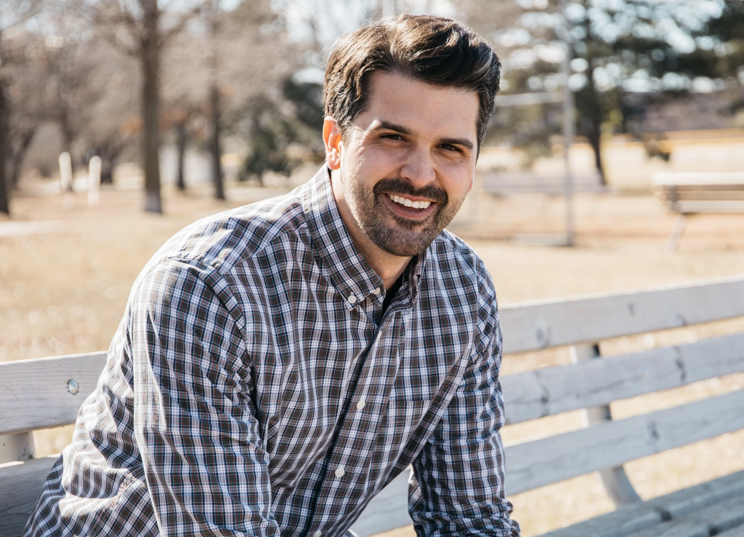 Man with dark hair and a beard smiling, wearing a plaid shirt, sitting on a park bench during daytime.