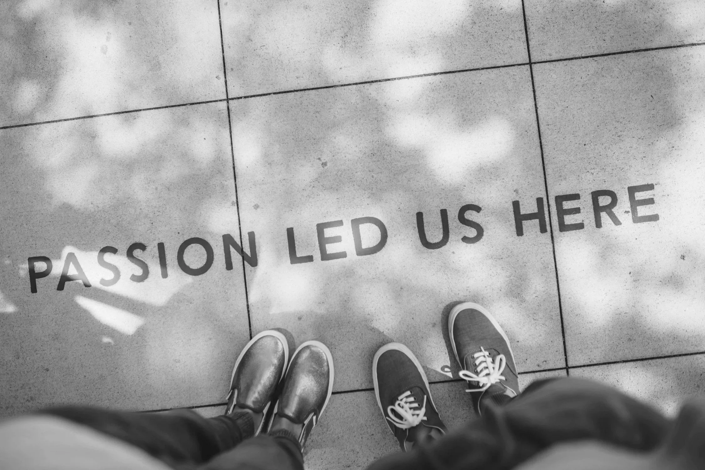 Black-and-white photo of two pairs of shoes standing on pavement with the words “Passion led us here” visible beneath them.