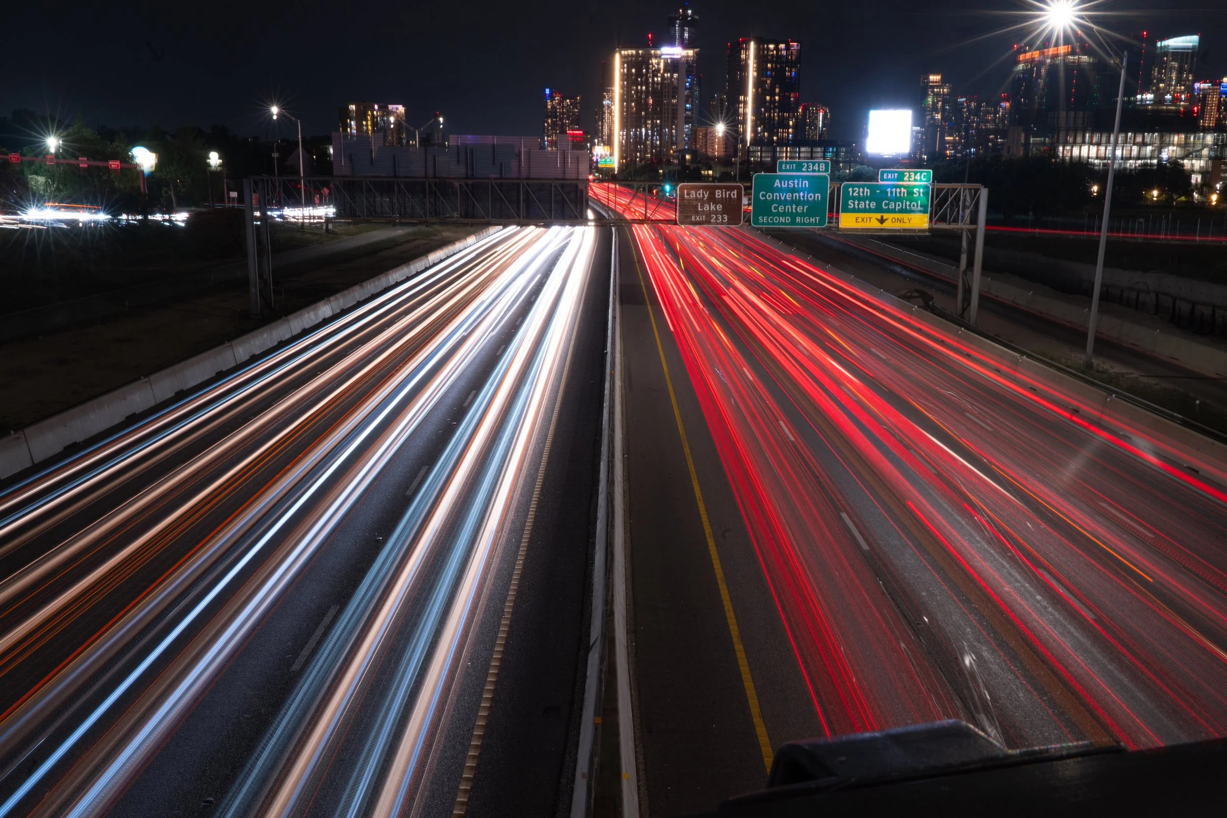 Long exposure photograph of a busy highway at night in Austin, Texas, with streaks of white and red lights from moving vehicles, illuminated city skyline in the background, and highway signs indicating exits to Lady Bird Lake, Austin Convention Center, and the State Capitol.