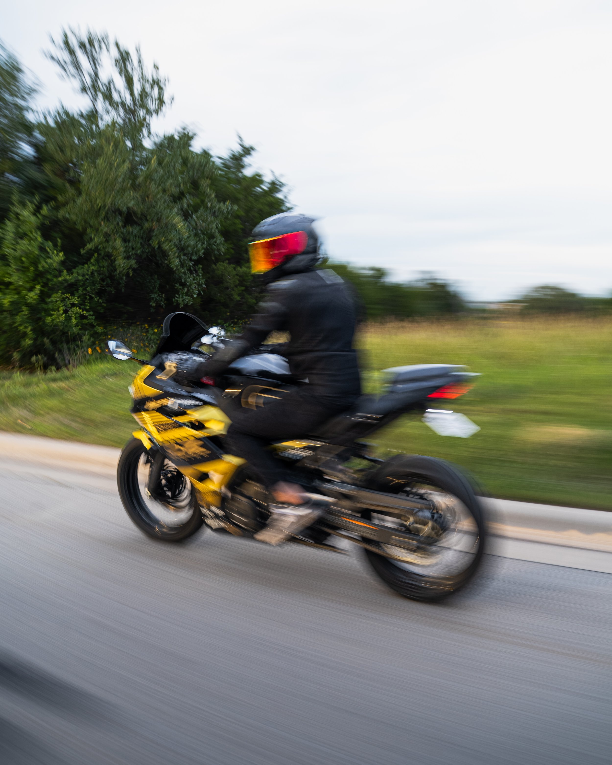 Blurry photo of a motorcyclist wearing a helmet, riding a yellow and black motorcycle on a road with green trees and open sky in the background.