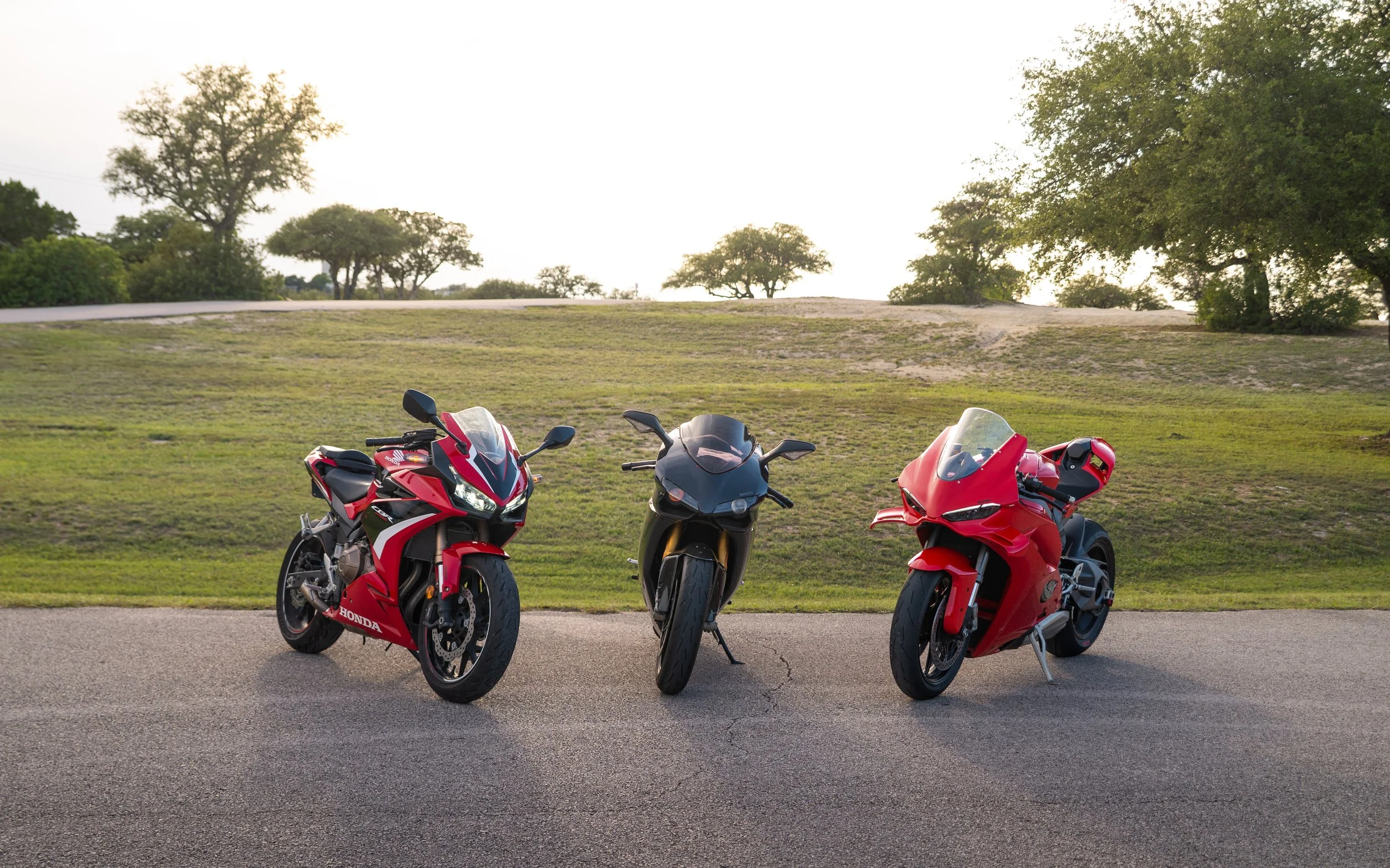 Three red and black motorcycles parked on the side of a road with green grass and trees in the background.