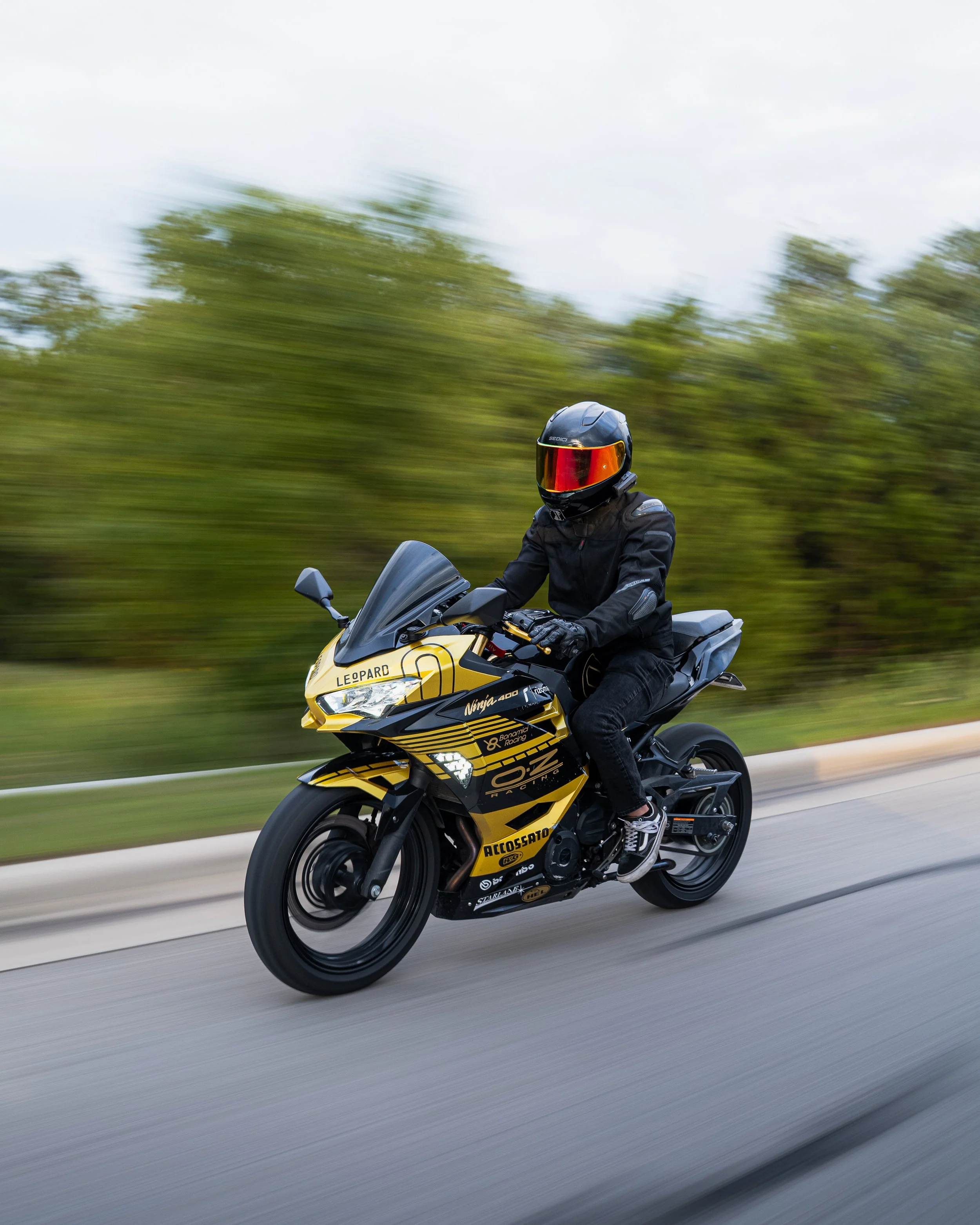 Motorcycle rider wearing black helmet and jacket riding a black and yellow sportbike on a highway, with blurred green trees and sky in background.