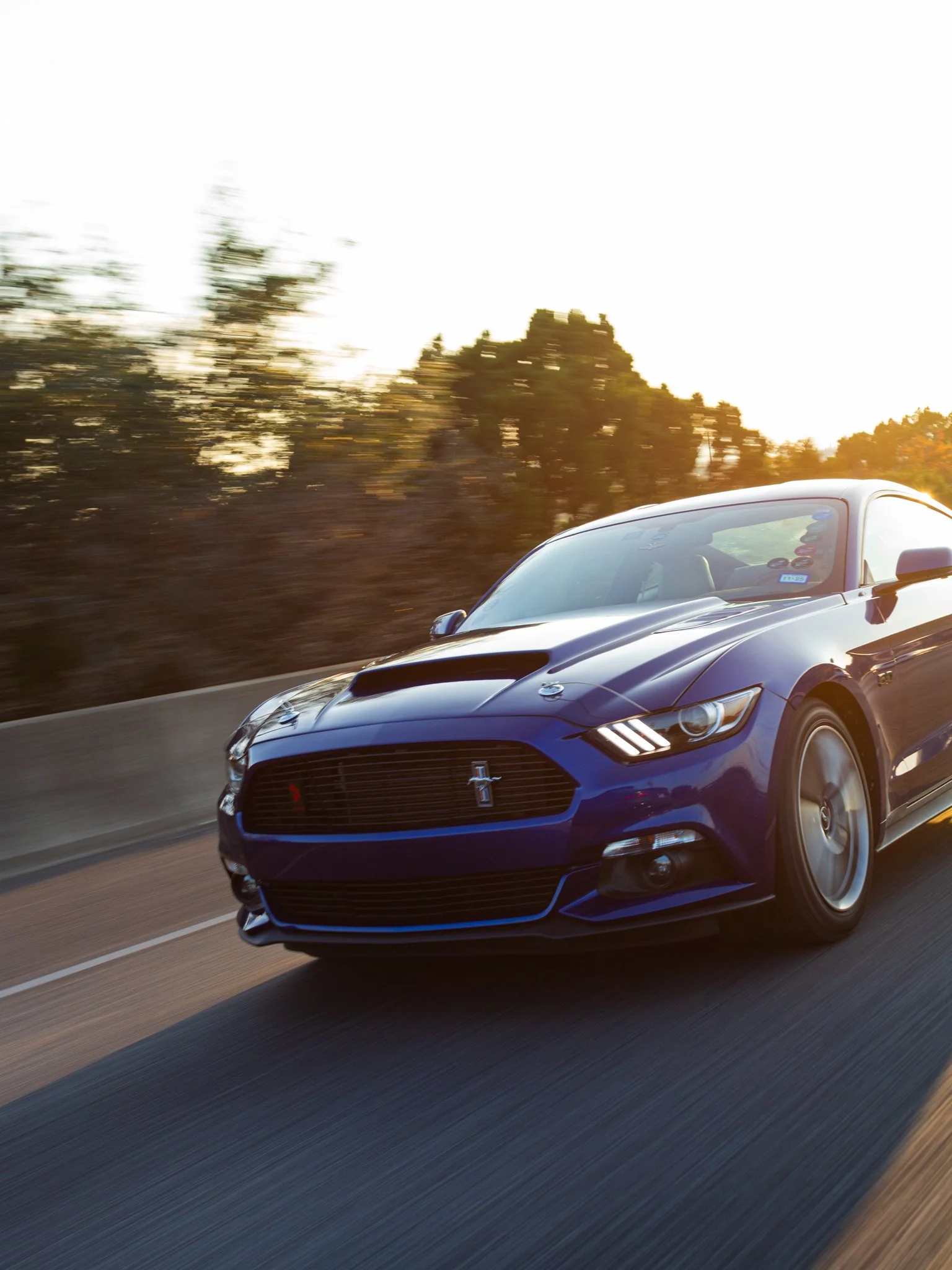 A blue Ford Mustang sports car driving on a highway with a blurred background of trees and sunset sky.