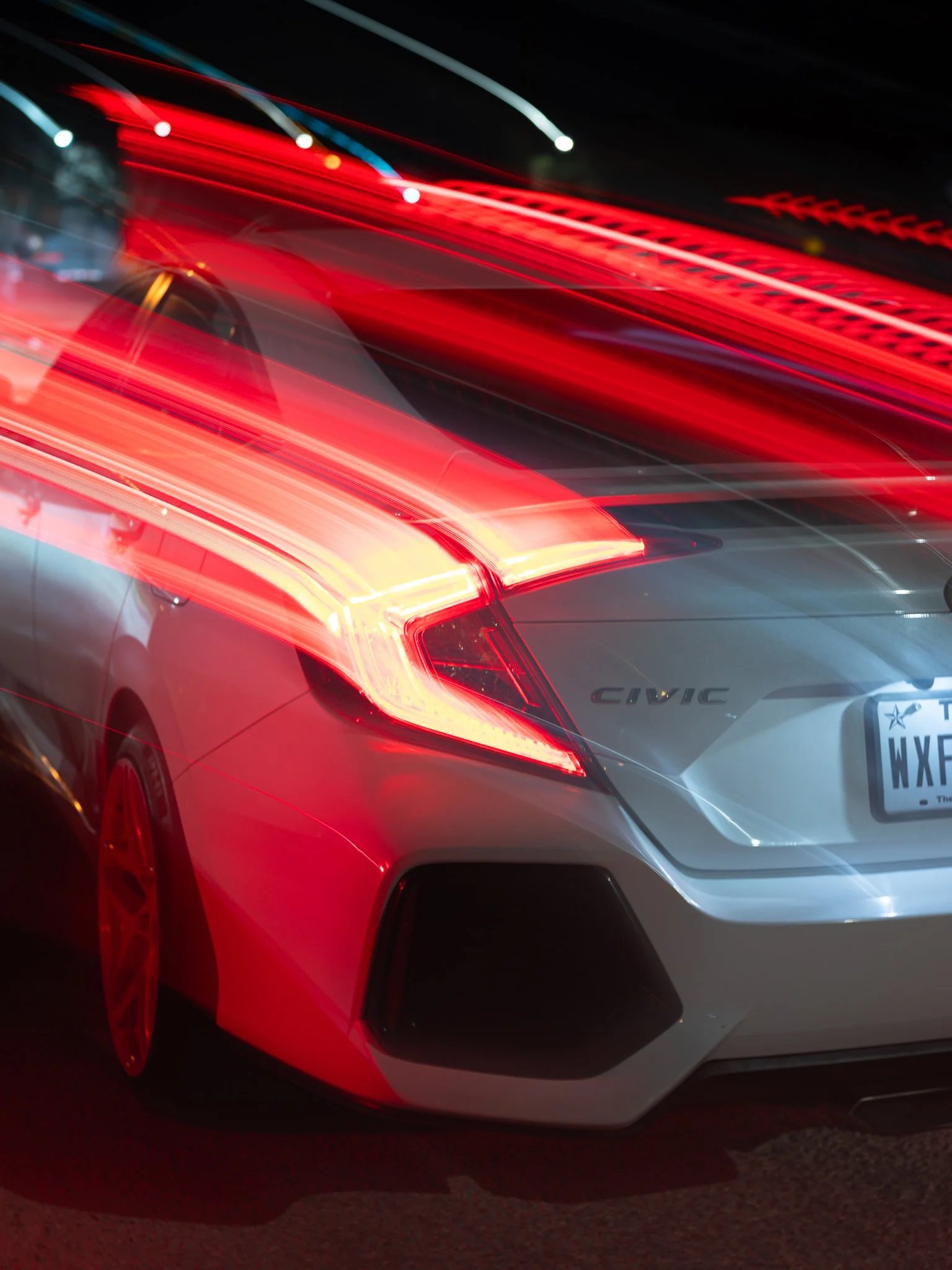 Close-up of a silver Honda Civic with illuminated red and white taillights, captured at night with light trails and motion blur.