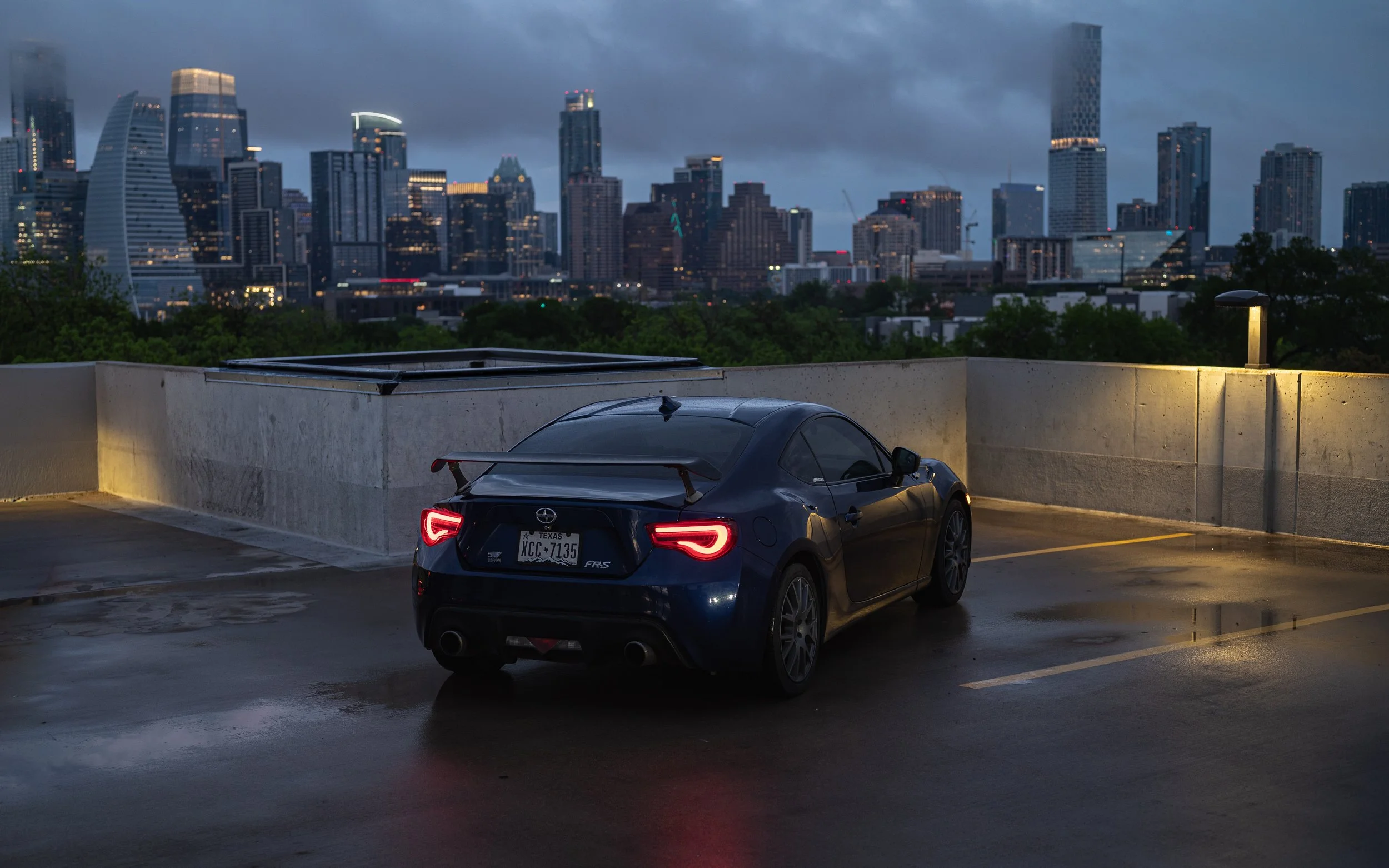A black sports car parked in a rooftop parking lot overlooking a city skyline at dusk. The city skyline features tall buildings with illuminated windows and a cloudy sky.