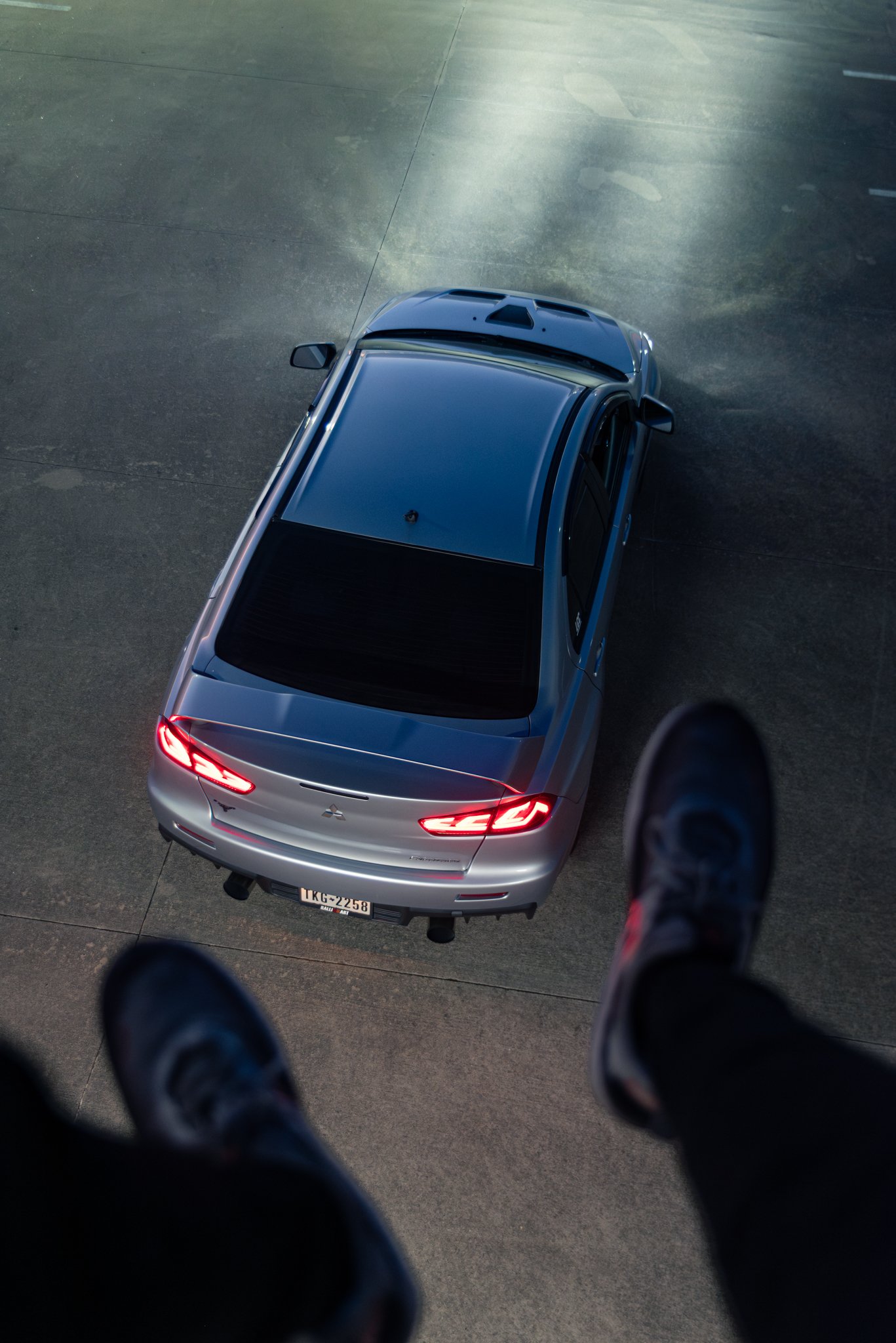 View from above of a silver car with tail lights on, on a paved surface, with a person's sneaker-clad feet visible in the foreground.