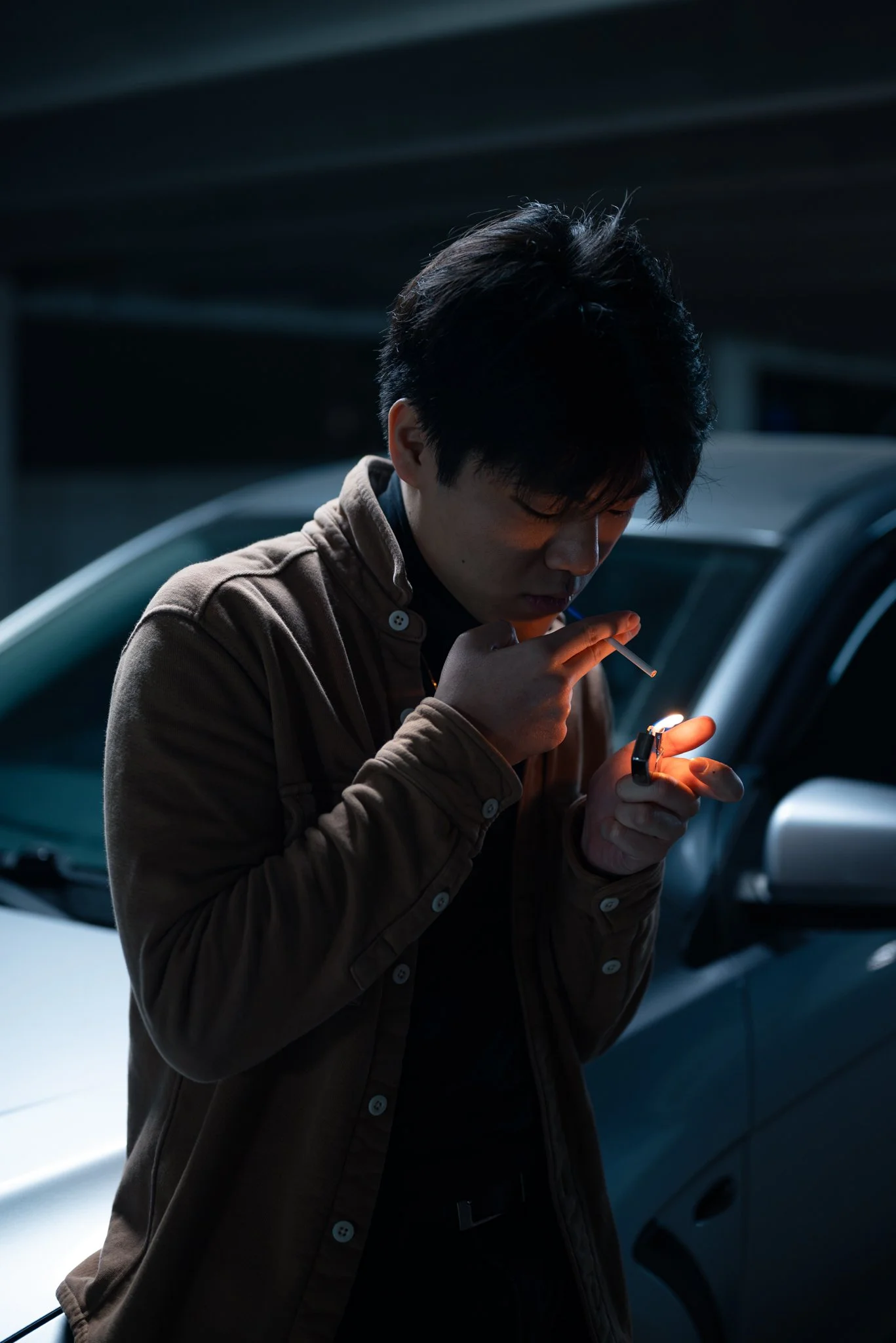 A young man with dark hair wearing a brown jacket, holding a lighter and a cigarette, standing next to a car in a dimly lit environment.