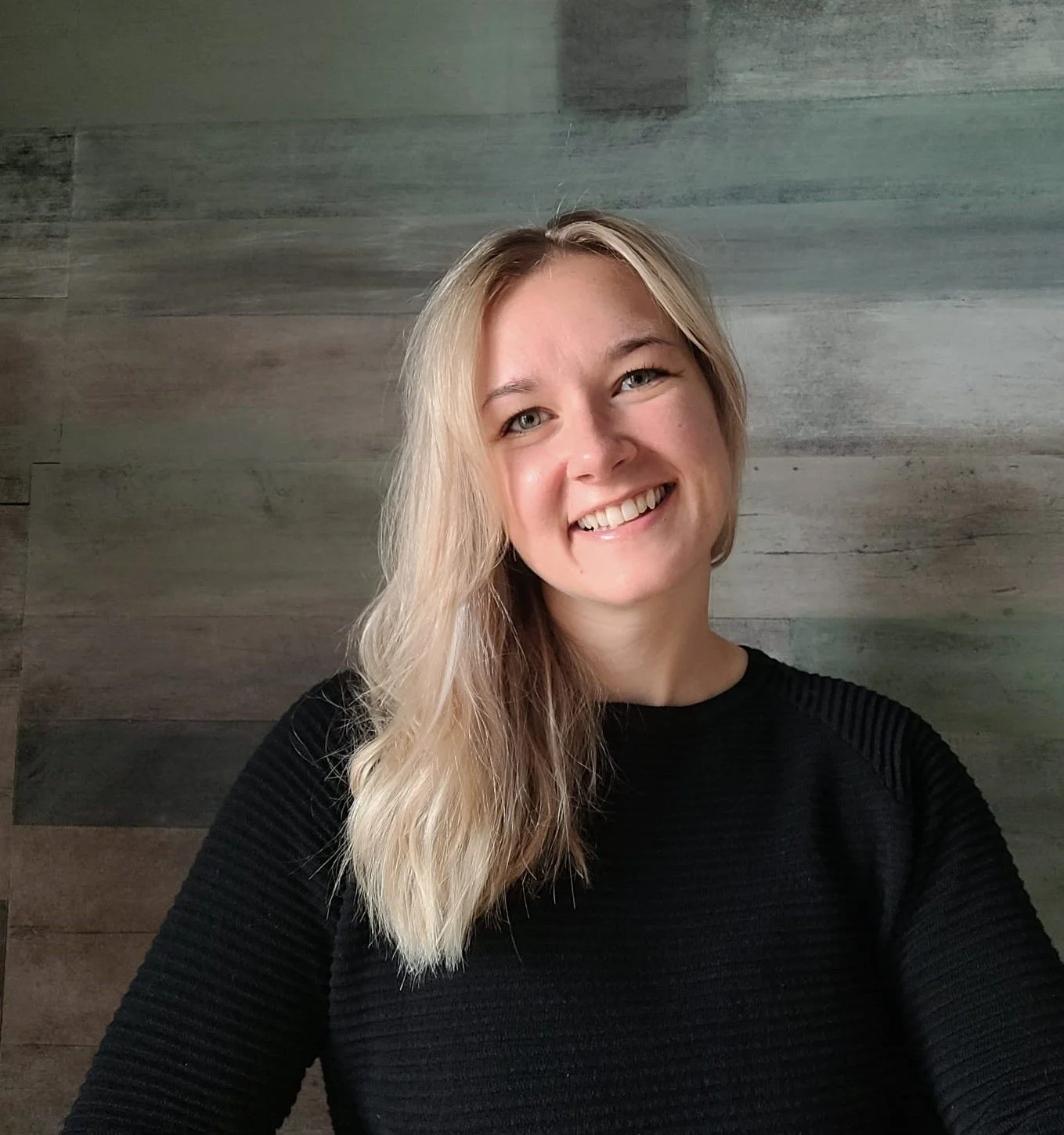 A woman with blonde hair smiling at the camera, wearing a black sweater, with a wood-paneled wall in the background.