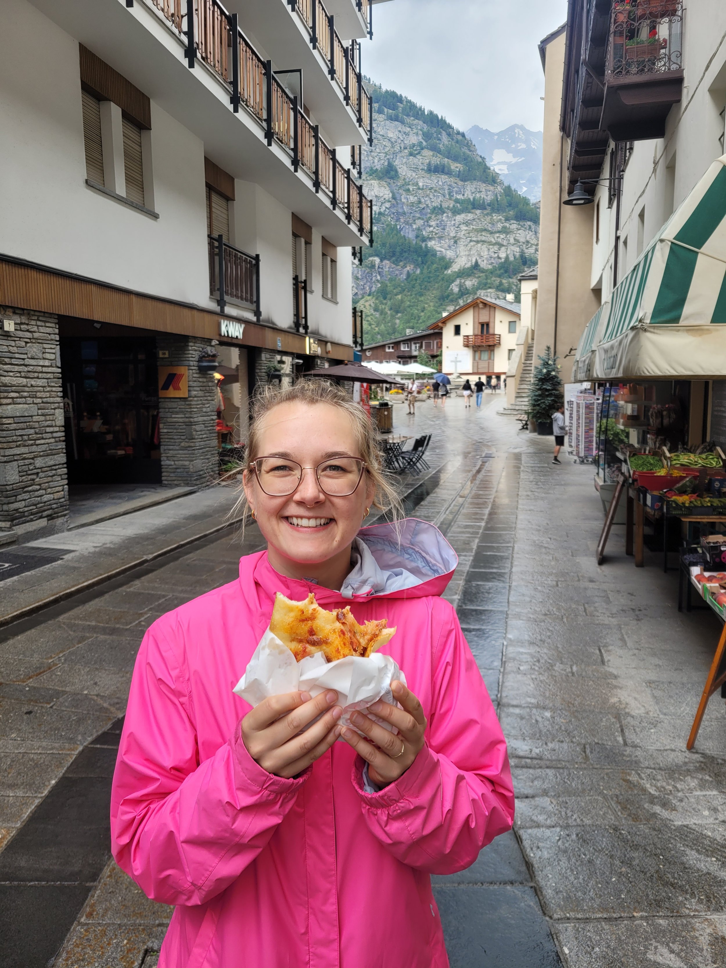 A smiling young woman wearing glasses and a pink jacket, holding a slice of pizza wrapped in paper, standing on a wet street in a mountain town.