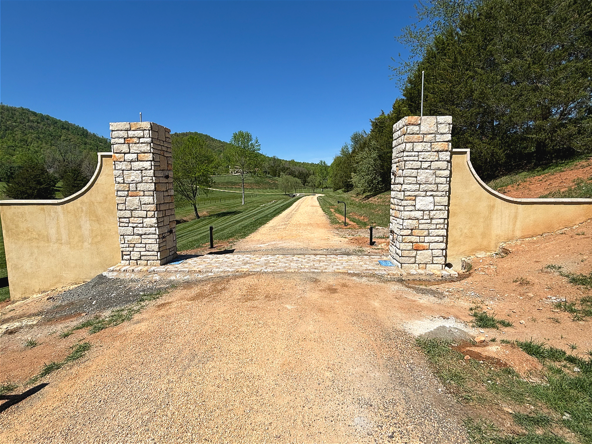 Entrance with stone pillars and a dirt road leading into a park or private property, surrounded by green trees and grassy hills under a clear blue sky.